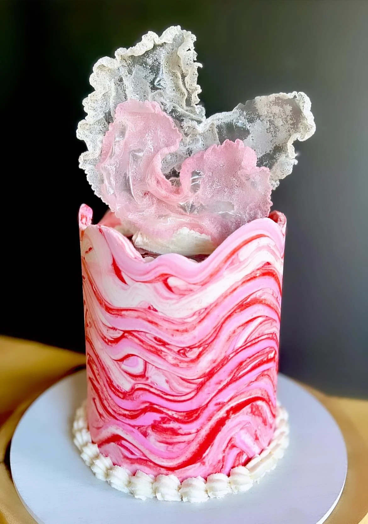 A tall, cylindrical cake with pink and white marbled icing, topped with a large, decorative pink and clear sugar lace made to resemble a flower.