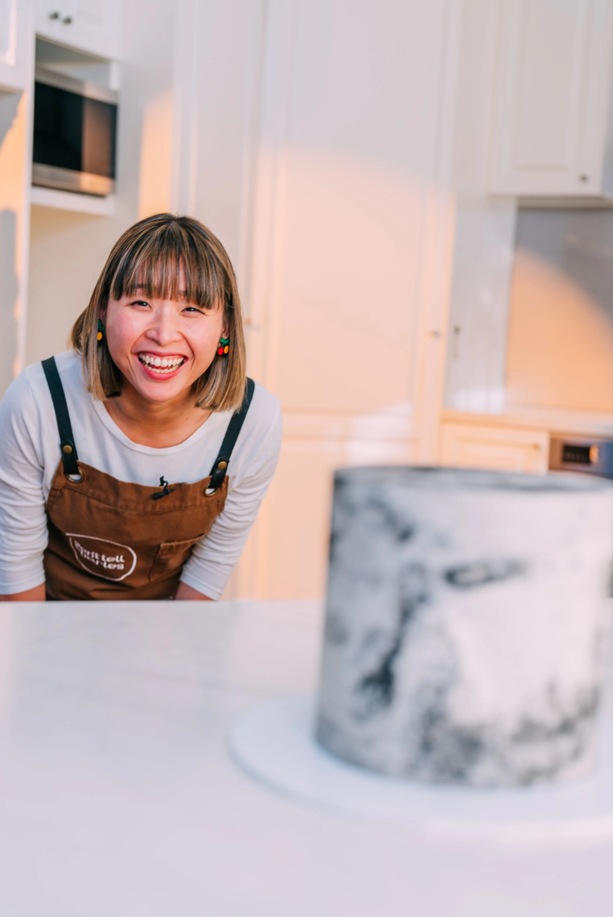 Woman with shoulder-length hair wearing cherries earrings, smiling happily, leaning forward in a kitchen with a white counter and a marble-patterned cake in the foreground.