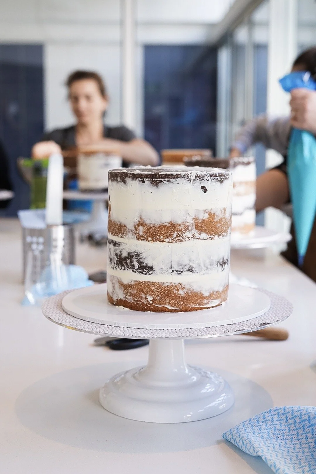 A partially decorated naked cake on a white cake stand, with layers of white frosting and visible sponge cake, sitting on a table in a bright room with people in the background.