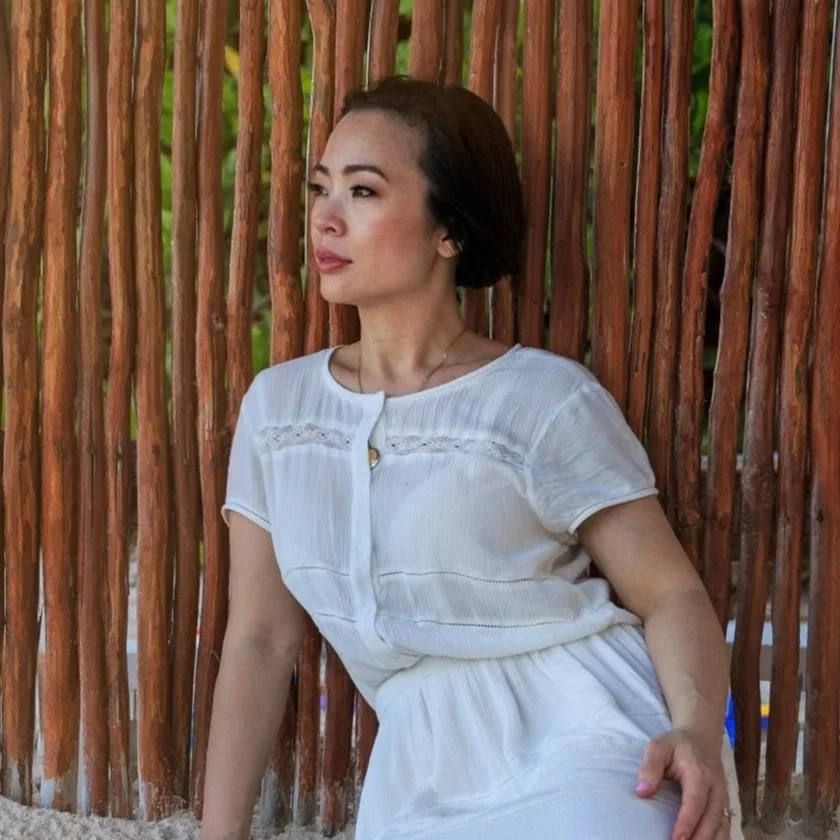A woman with short dark hair wearing a white dress, sitting against a rustic wooden fence made of vertical sticks, outdoors with green trees in the background.