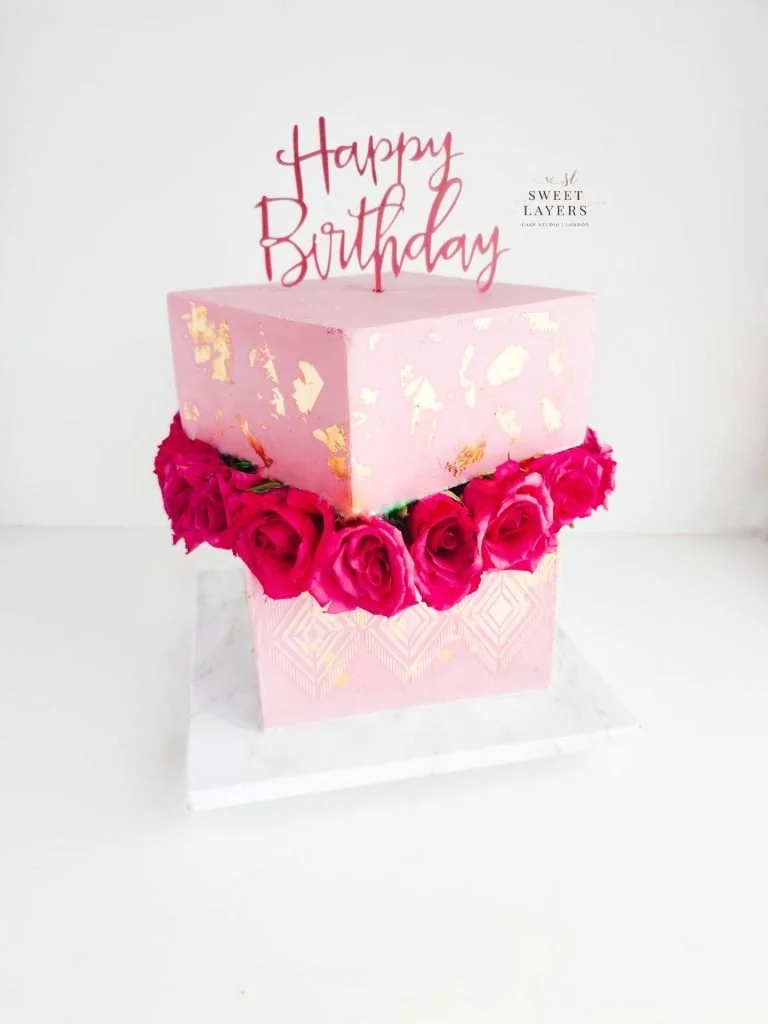A pink birthday cake with gold leaf accents, decorated with pink roses around the base, with a topper that says "Happy Birthday" in red cursive writing.