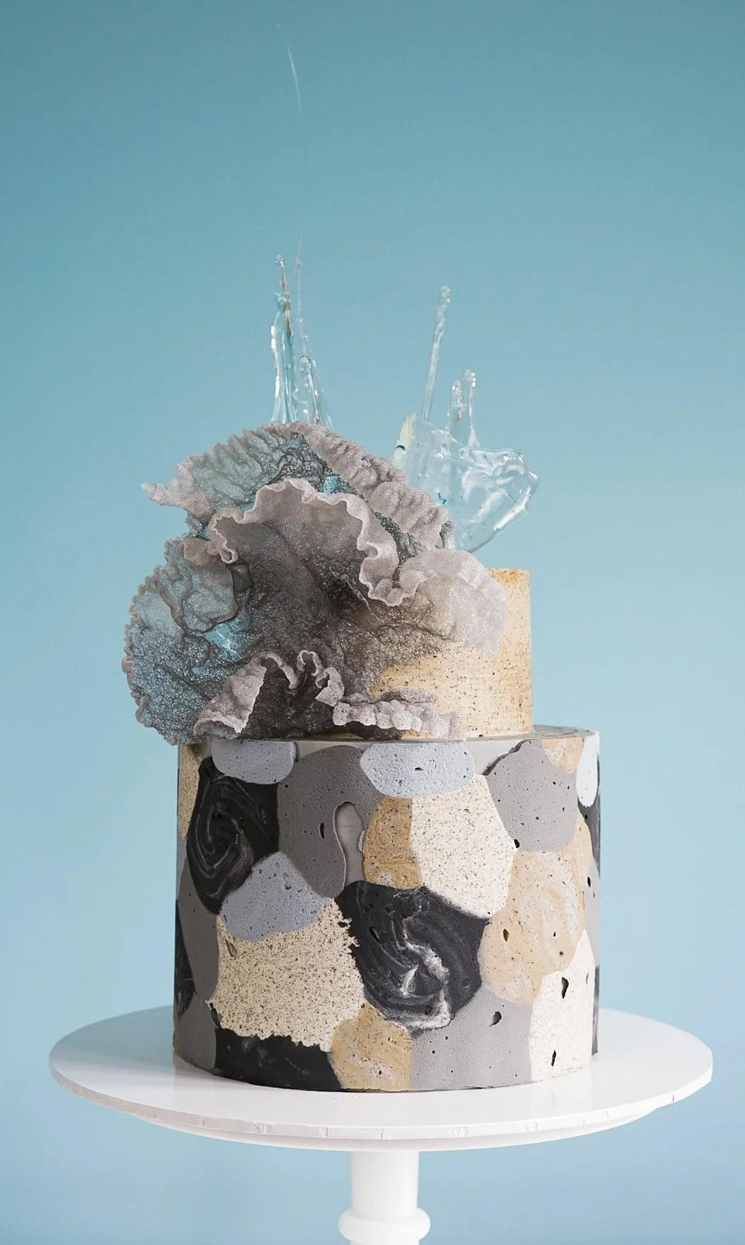 A modern, artistic cake with a abstract marbled black, white, gray, and beige design, topped with a decorative gray flower-like sugar sculpture and a clear glass-like decoration, set against a light blue background.