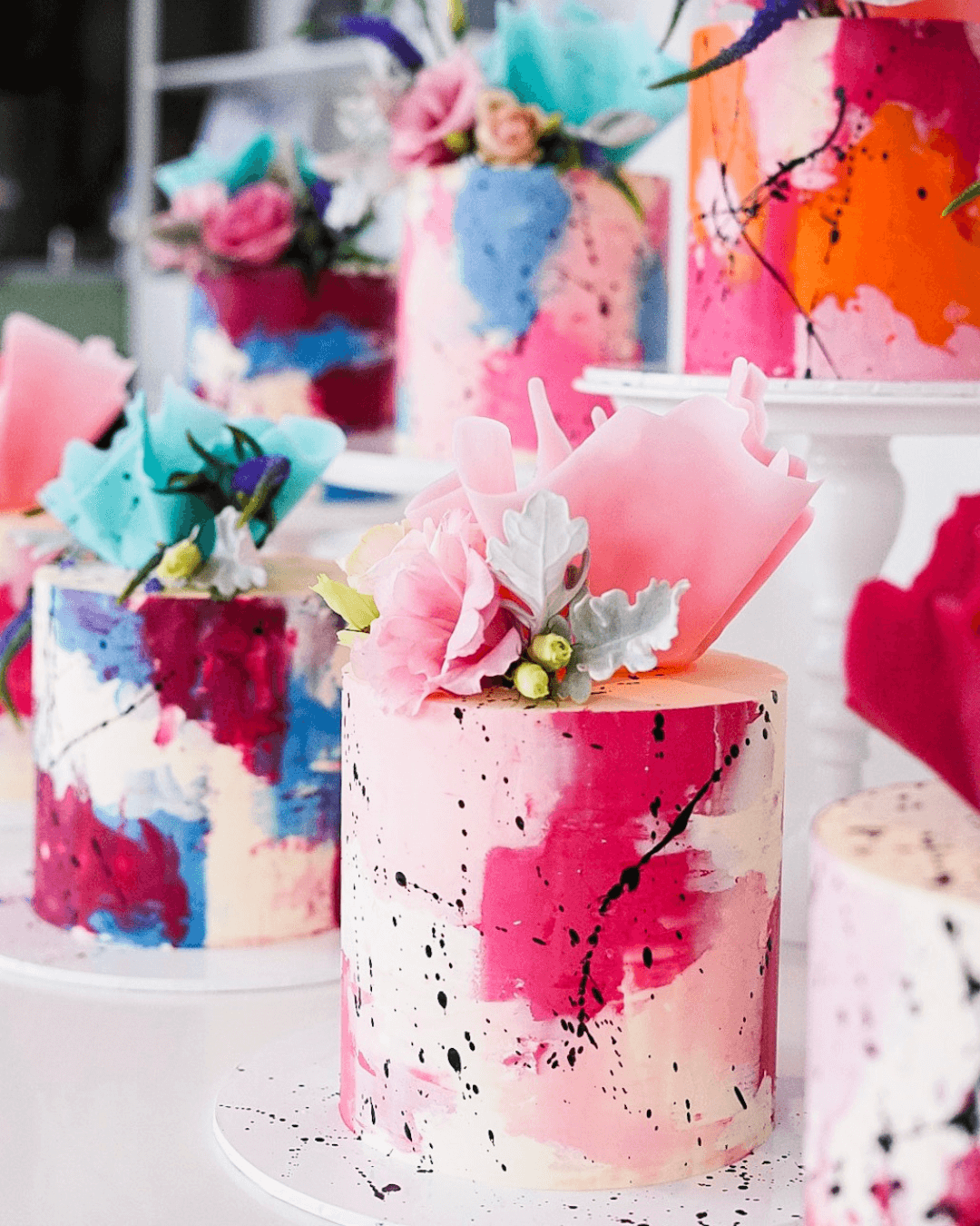 Colorful abstract cakes with floral and splatter paint decorations displayed on a table.