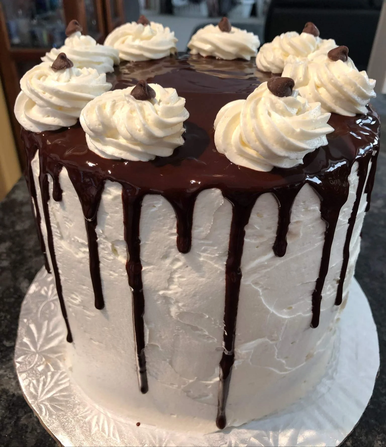 A tall white frosted cake with chocolate ganache drips on the sides, topped with whipped cream swirls and small chocolate chips, placed on a decorative silver cake board.