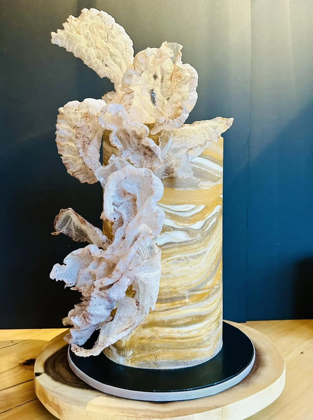 A decorative arrangement featuring a cylindrical marble vase with a swirling pattern in shades of yellow, white, and beige, containing ruffled light-colored dried or artificial leaves or coral on a black round base, set on a wooden surface against a dark background.