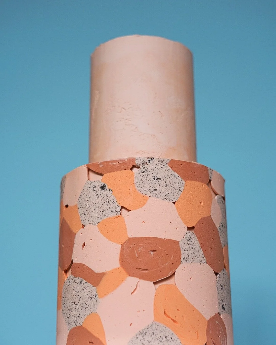 Close-up of two cylindrical objects with pastel colors against a blue background. The lower object features a terrazzo pattern with various circular fragments in shades of peach, beige, and gray. The upper object is a solid light pink color.