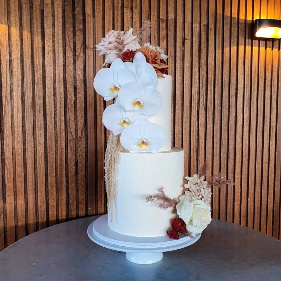 A two-tier white wedding cake decorated with white orchids, roses, and dried flowers, set against a wooden backdrop.