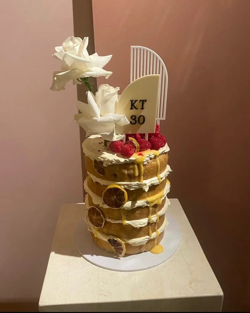 A tall, multi-layered cake decorated with white roses, raspberries, lemon slices, and a white decorative topper with the text 'KT 30'.