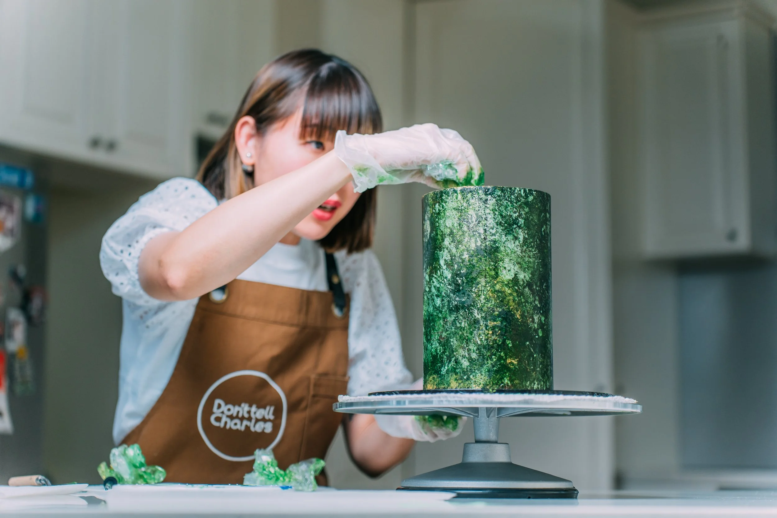 A woman wearing a brown apron and gloves decorates a green galaxy-themed cake in a kitchen.