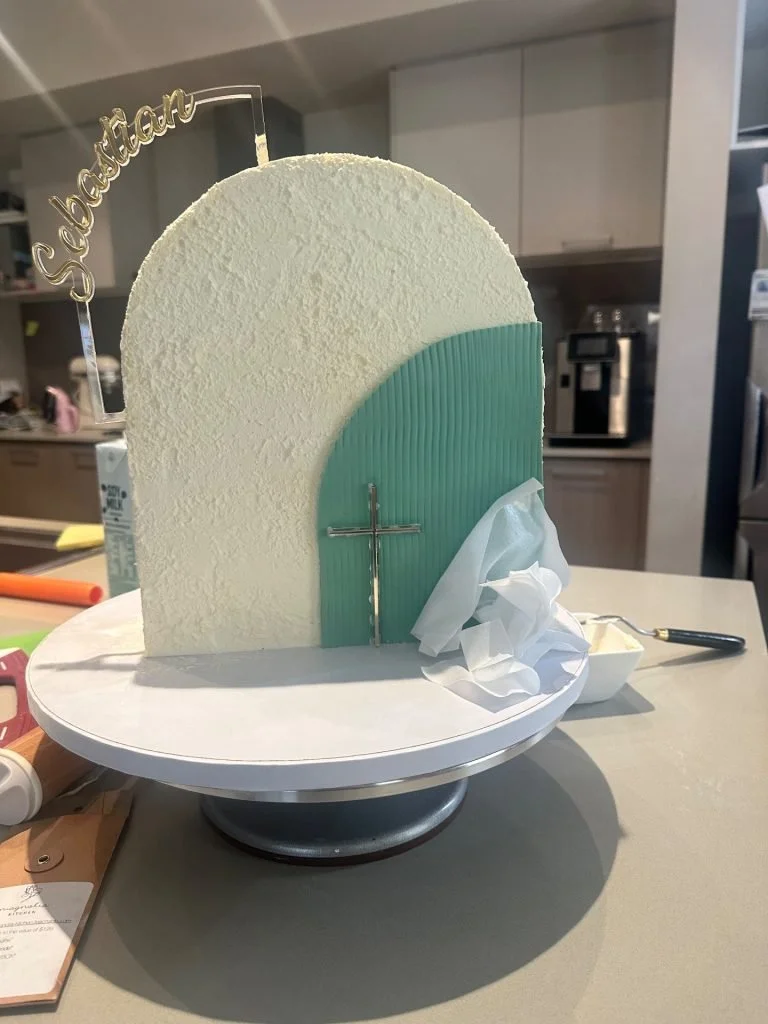 A decorated cake with a white textured surface, a light blue semi-circular panel, a silver cross, a tissue, and a cake topper that says 'Salutations' in gold. The cake is on a round white base on a table.
