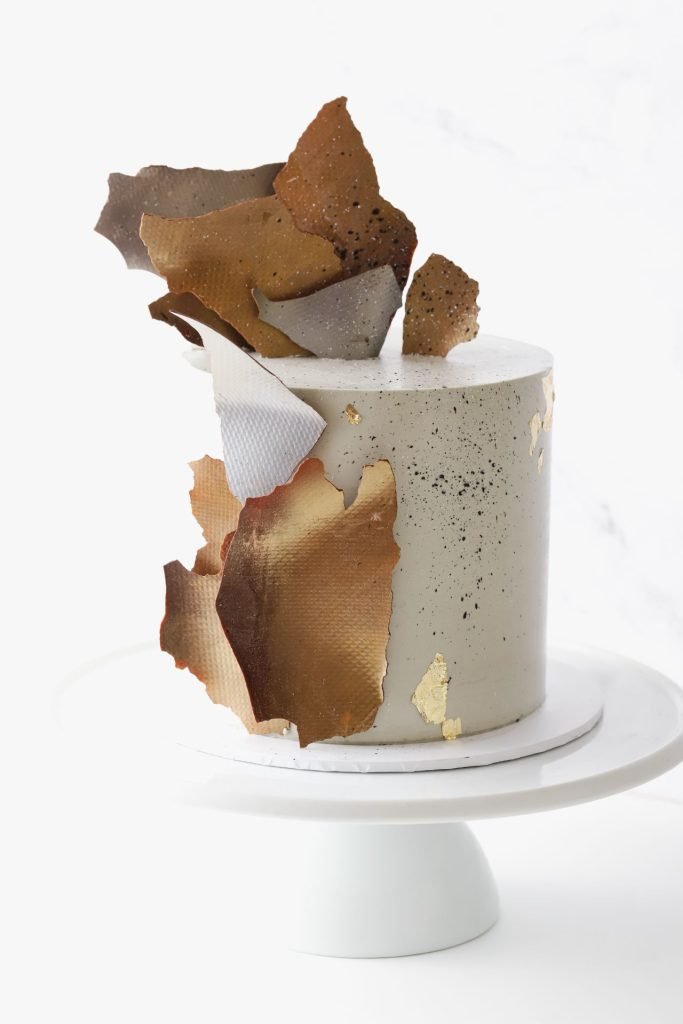 Gray frosted cake decorated with gold and metallic leaf accents, topped with irregular-shaped chocolate or caramel shards, on a white cake stand against a white background.