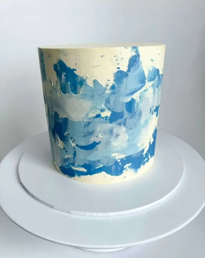 A round cake with a marbled blue and white design on its surface, placed on a white cake board.