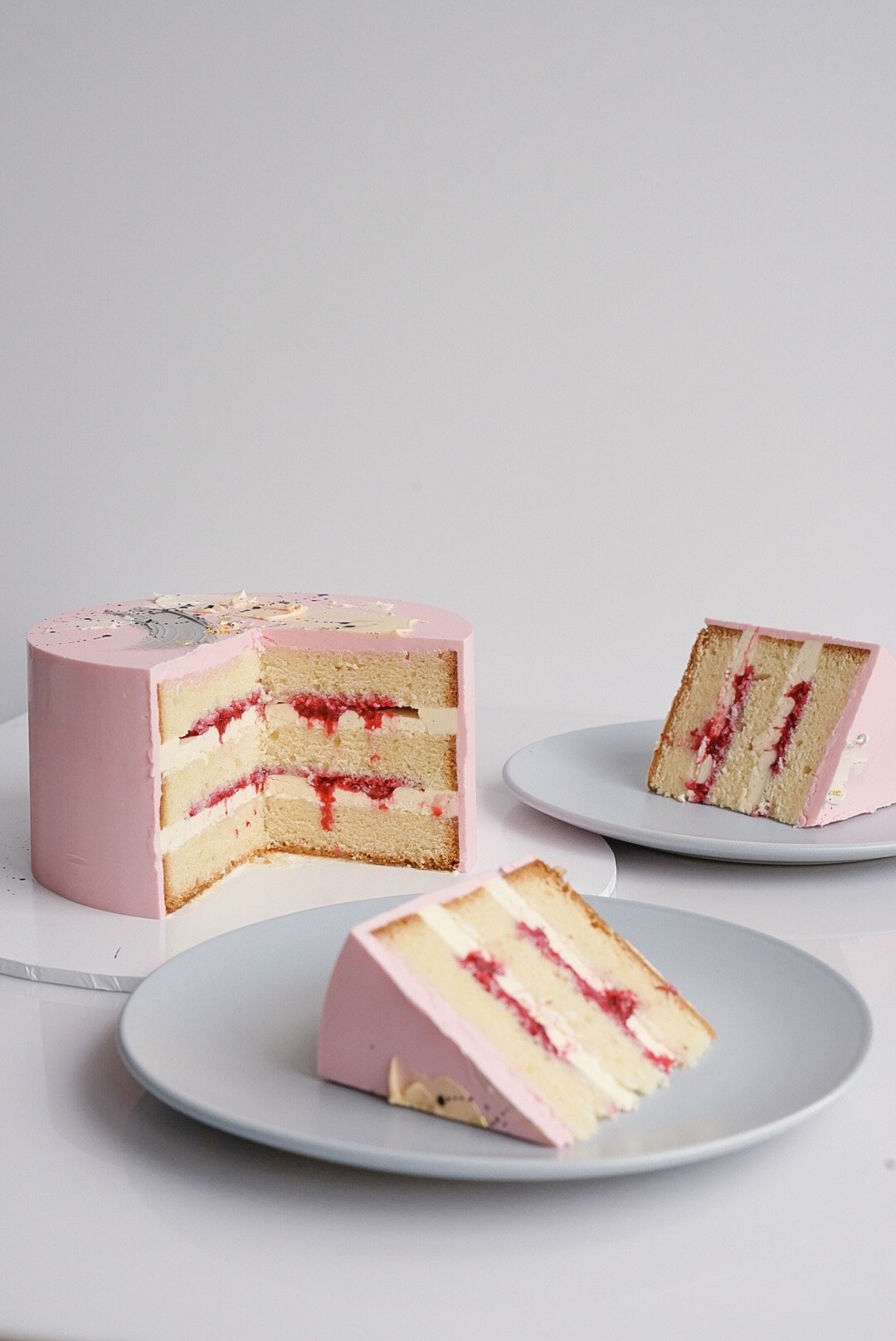 A pink layered cake with white frosting and red filling, with one slice on a white plate and the rest on a cake stand, all set against a plain white background.