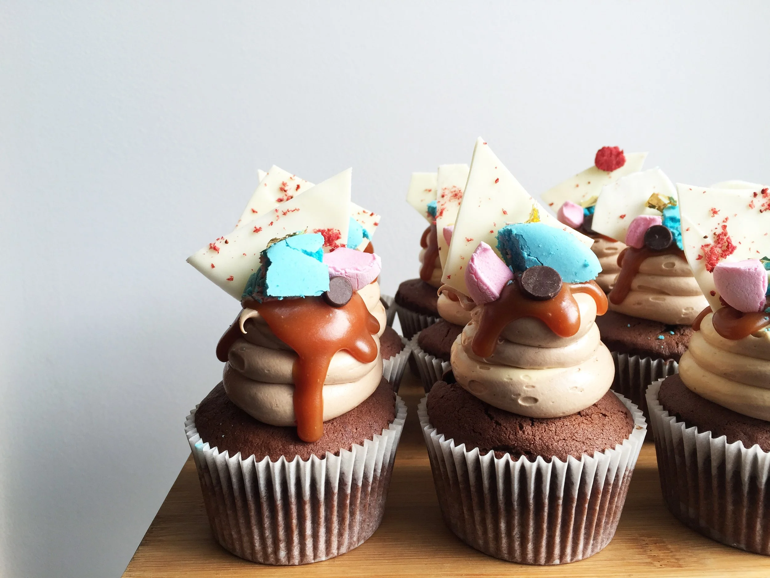 Chocolate cupcakes decorated with swirled frosting, caramel drizzle, and colorful candy pieces topped with white chocolate shards on a wooden surface.