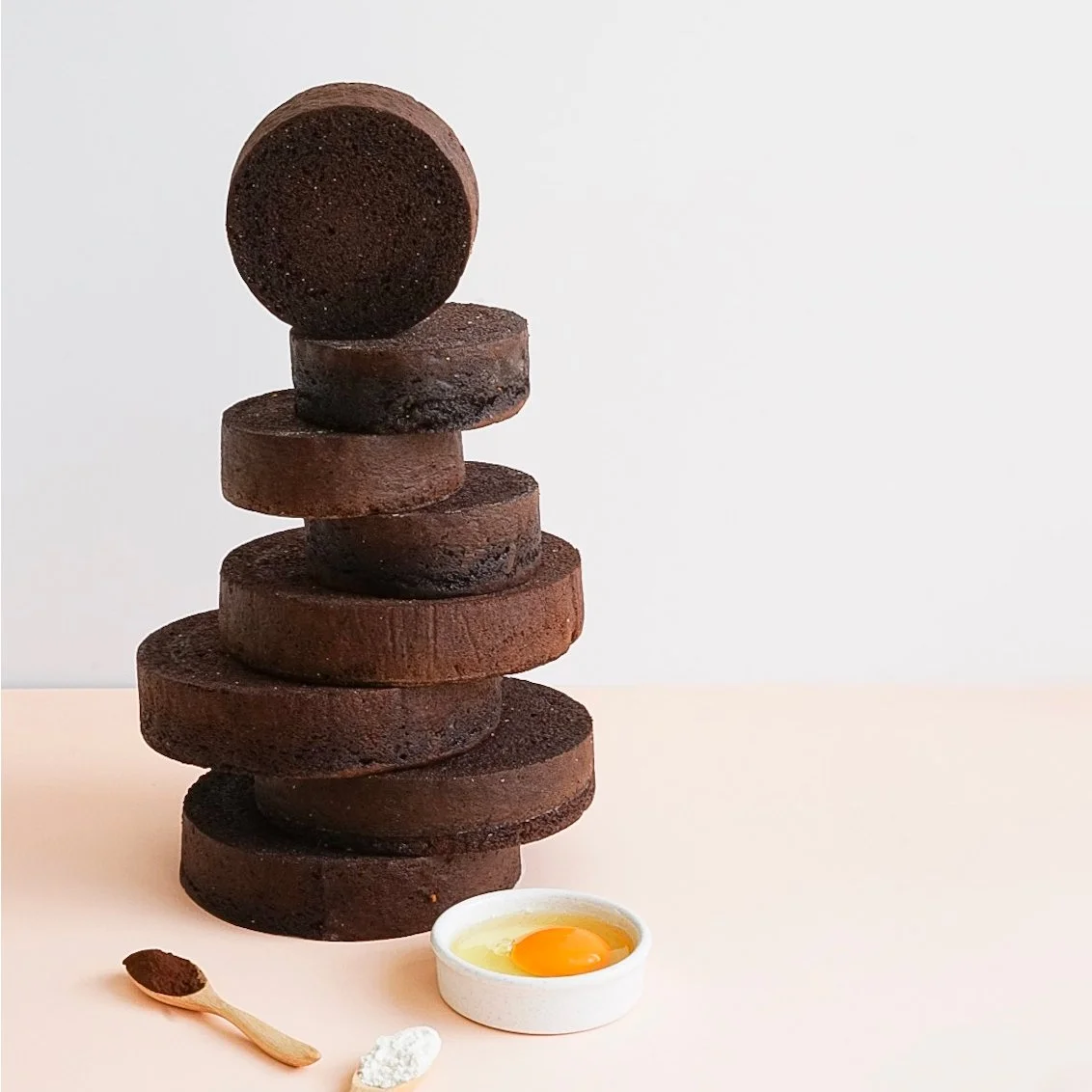 Stacked chocolate cookies with a raw egg in a small dish, a wooden spoon with cocoa powder, and a white scoop of flour on a pink surface.