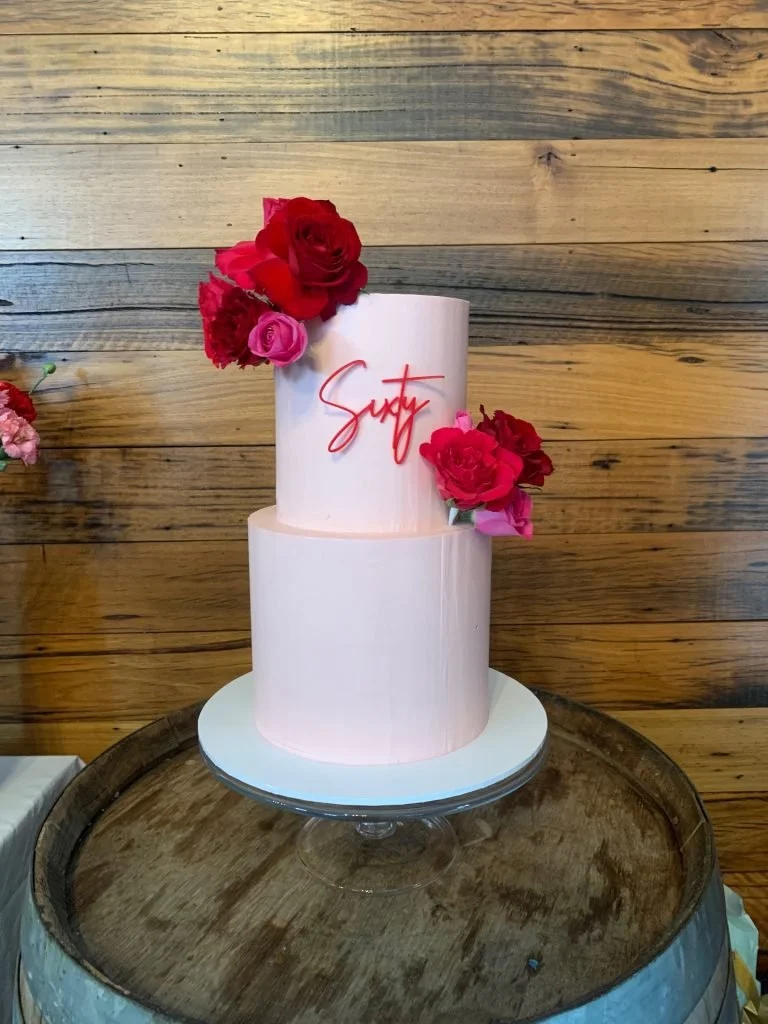 A two-tier pink cake decorated with red and pink flowers, with the word 'Savy' written in red on the top tier, displayed on a glass cake stand on a wooden barrel against a wooden wall background.