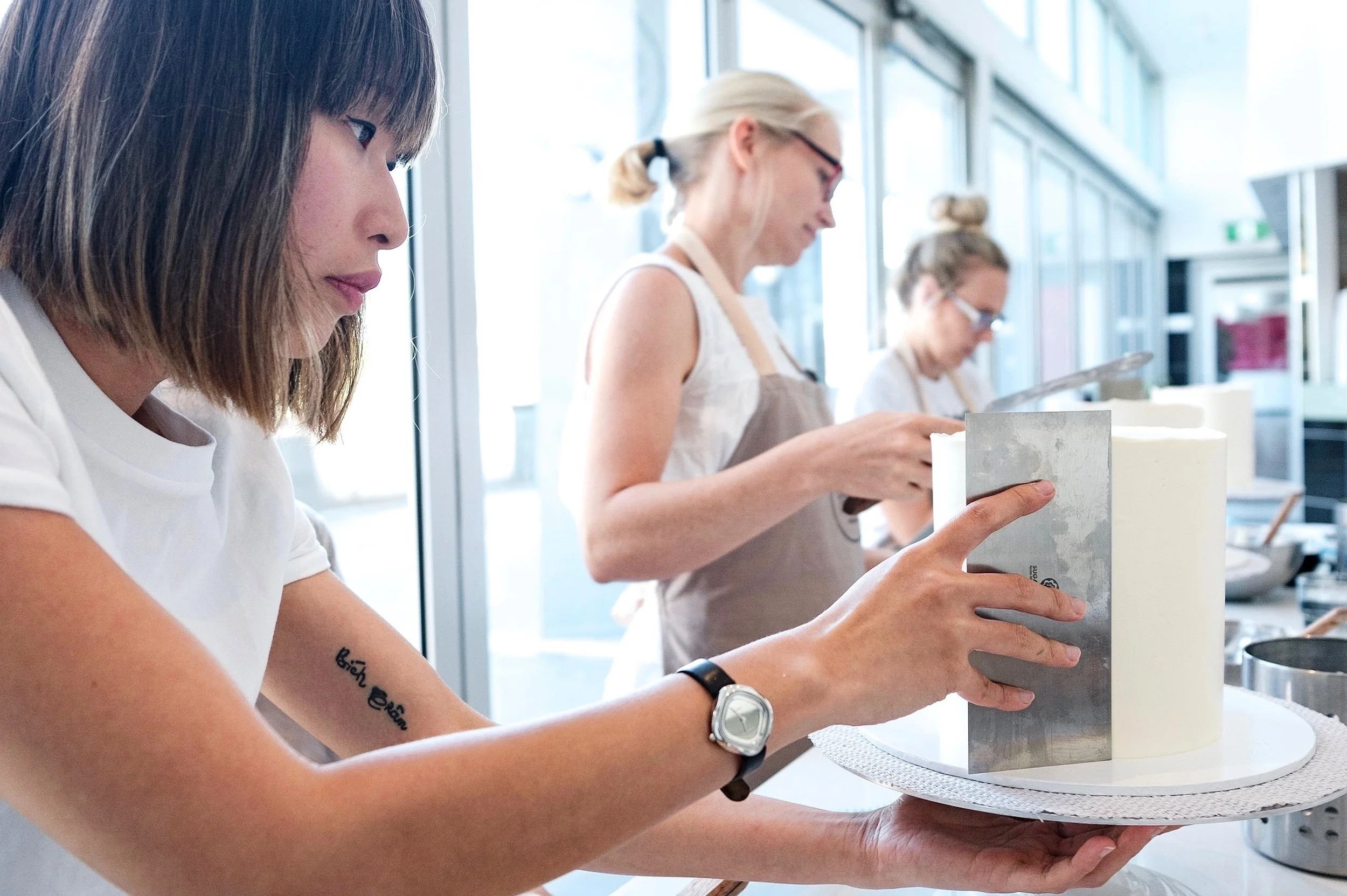 A woman decorating a large, white, round cake with a metal tool in a bright kitchen environment.