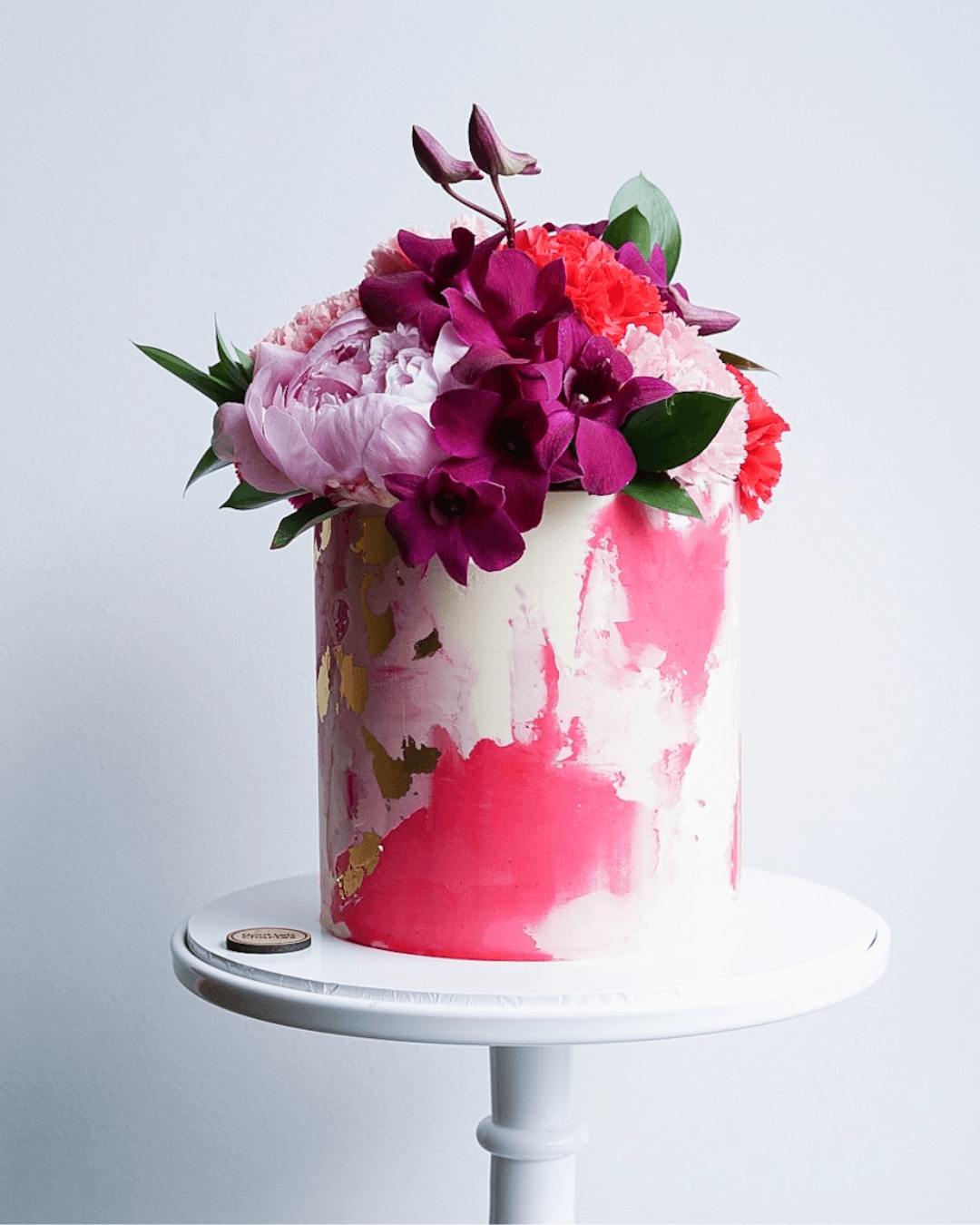 Pink and purple floral arrangement in a tall cake with watercolor pink and gold design on a white cake stand.