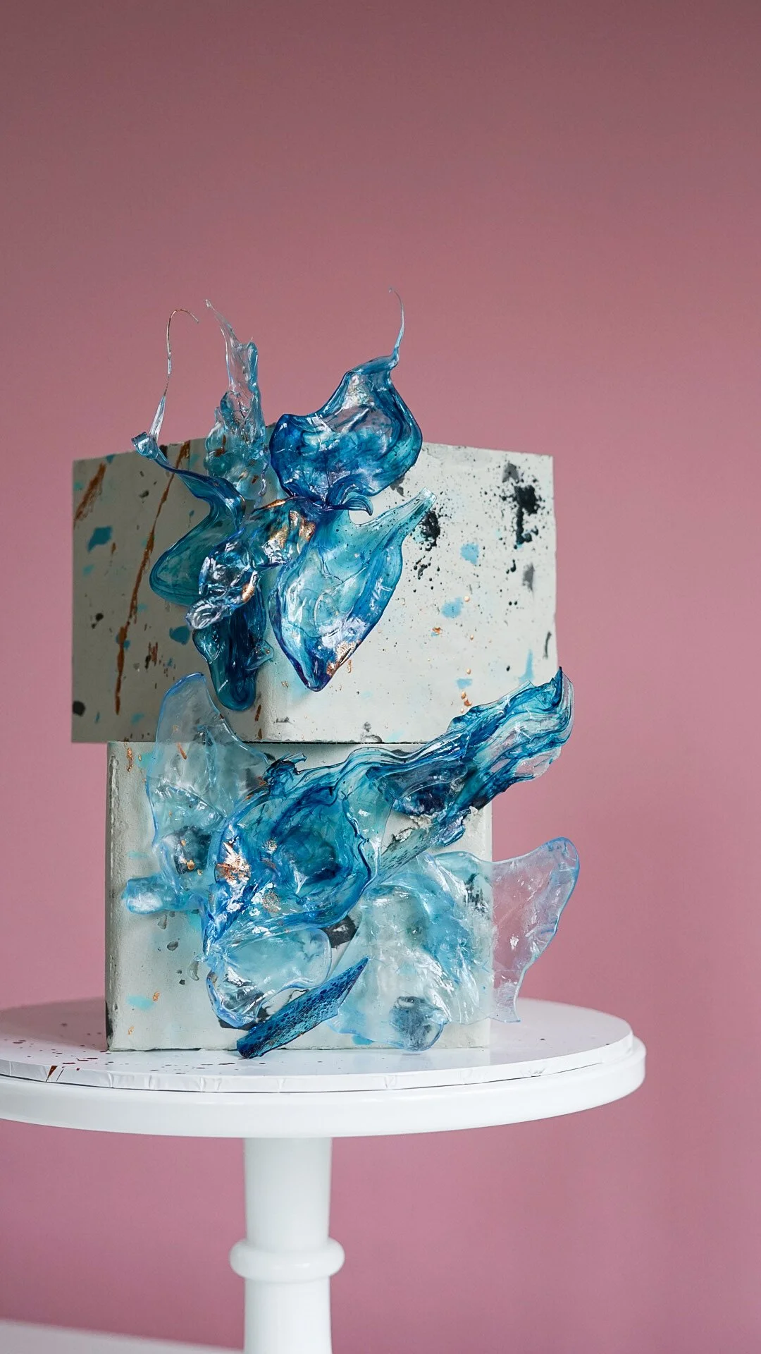 Glass art sculpture with abstract blue and clear glass pieces on a rectangular white and speckled pedestal, set against a pink background.