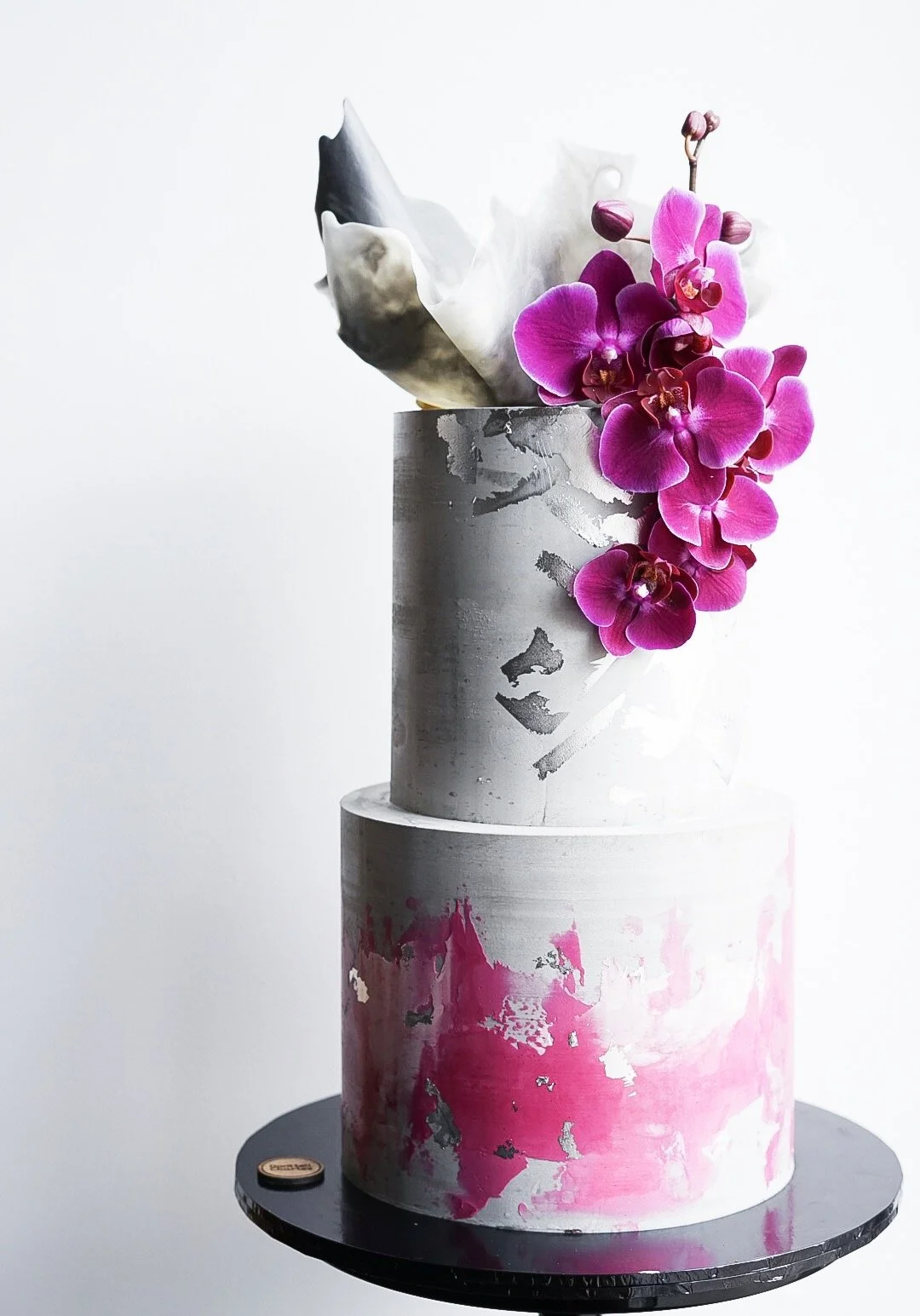 A two-tiered cake with a silver and pink paint splatter design, topped with pink orchids and abstract white and black decorations.