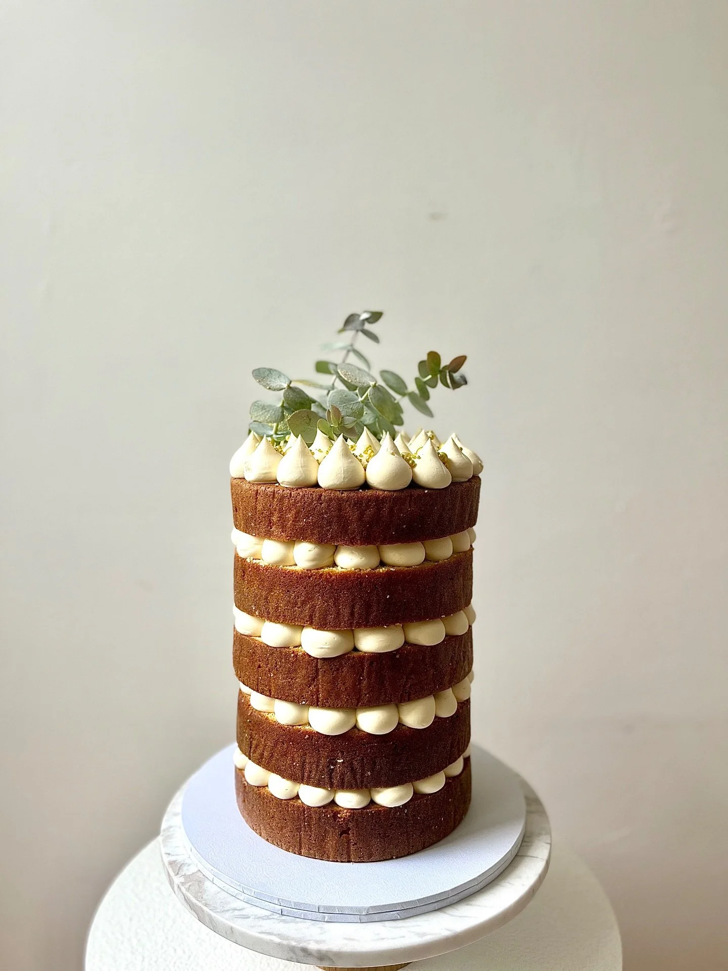 A tall, layered chocolate cake with cream filling and frosting, topped with green leaves and gold sprinkles, on a white cake stand.