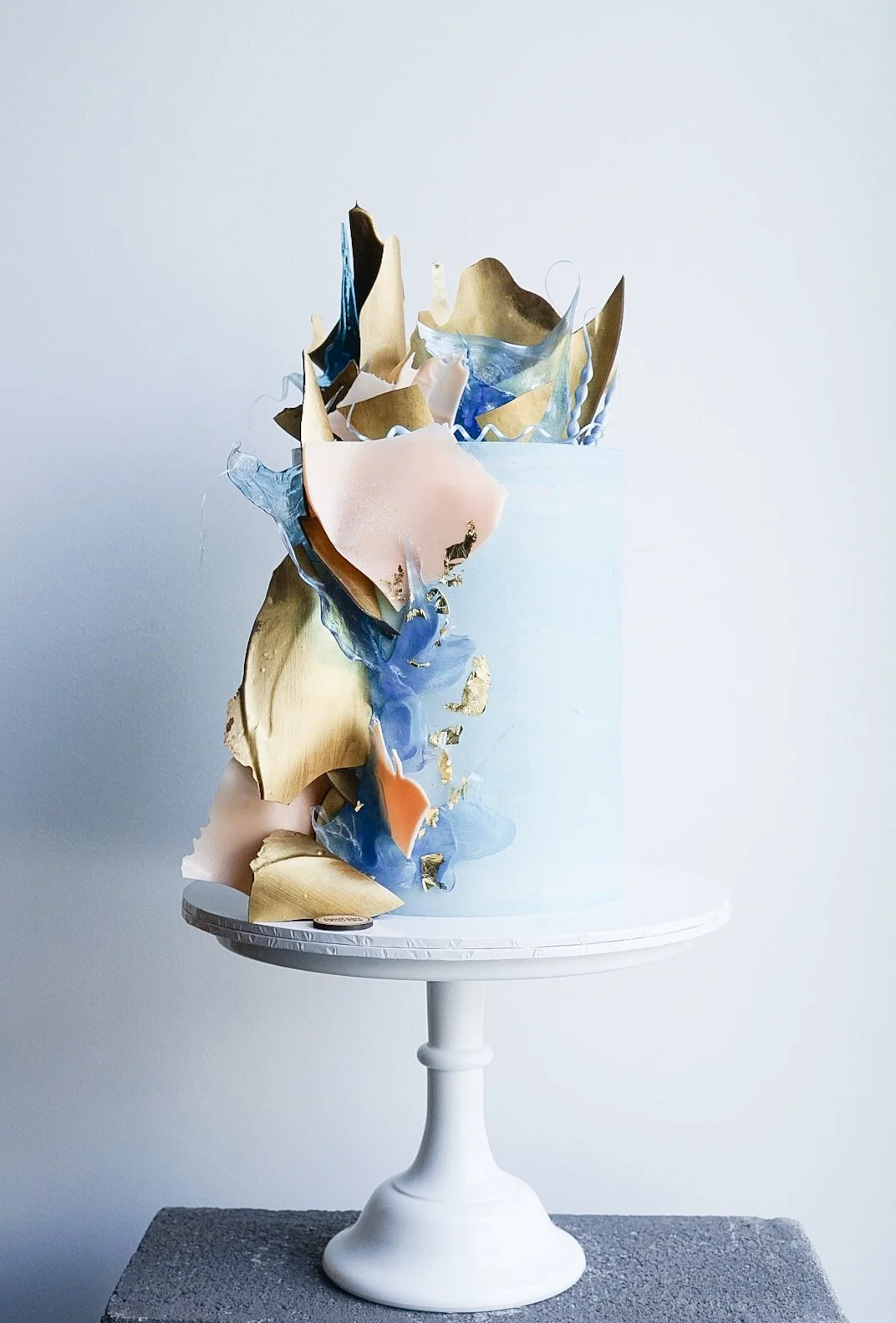 A tall cake with abstract, colorful, and metallic toppers on a white cake stand against a plain light wall.
