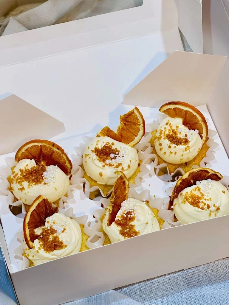 Six cupcakes with white frosting, topped with dried orange slices, white sprinkles, and crumbled brown topping, in a white box on a table.
