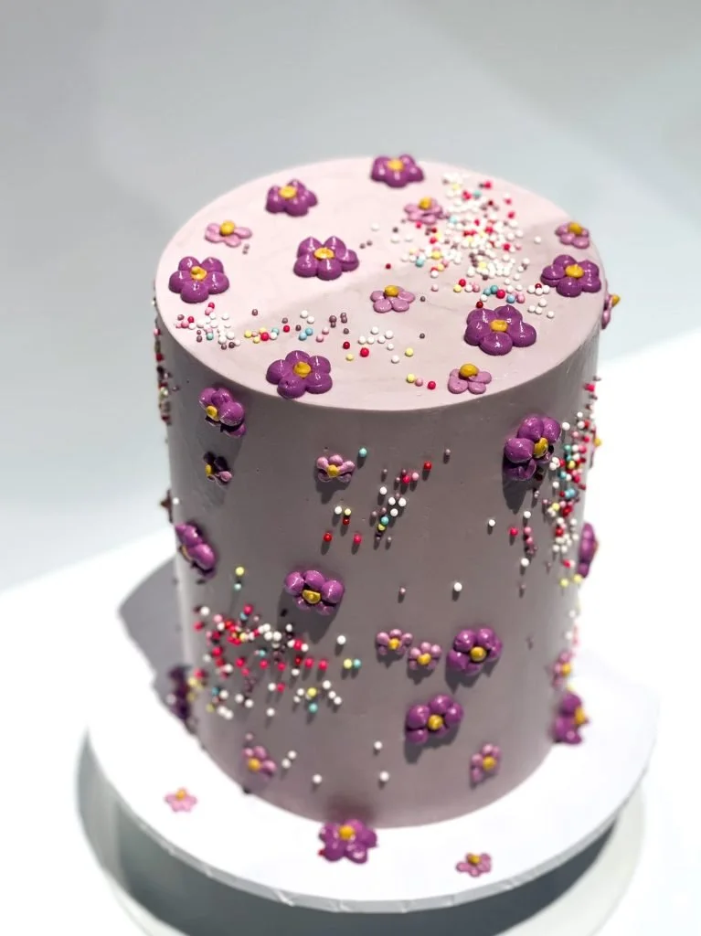 A purple cylindrical cake decorated with purple and yellow flower-shaped icing and multicolored sprinkles on top and sides.