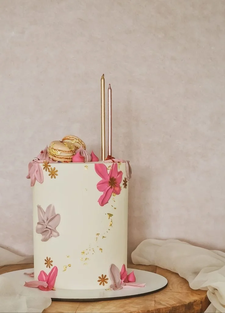 A tall, white birthday cake decorated with pink and beige flowers and gold accents, topped with macarons, pink frosting dollops, and two tall candles. The cake is on a round black-and-white base on a wooden surface, with white cloth around it.
