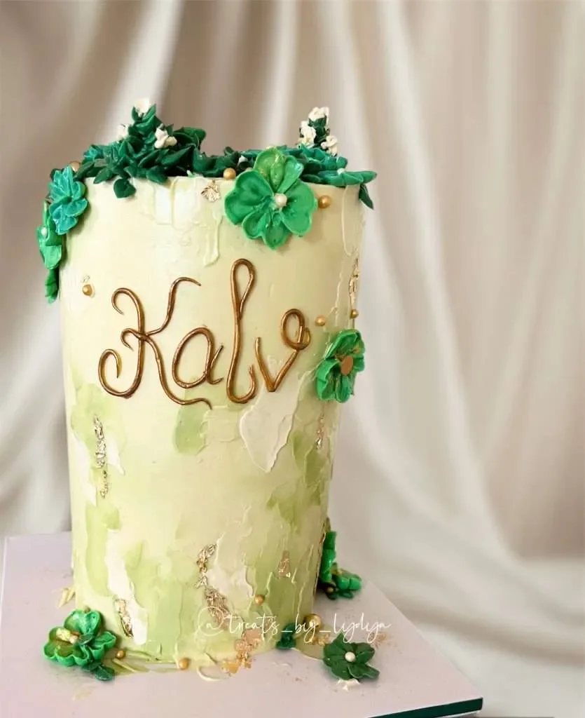 A tall, cylindrical cake decorated with green flowers and gold accents, with the name "Kale" written on it in gold lettering.