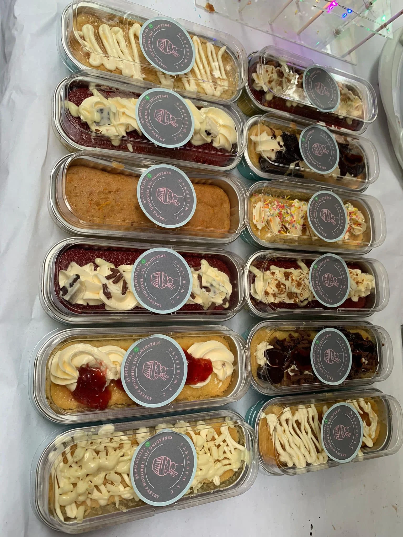 Various clear plastic containers of baked goods with labels, including cakes and pastries, arranged on a table.