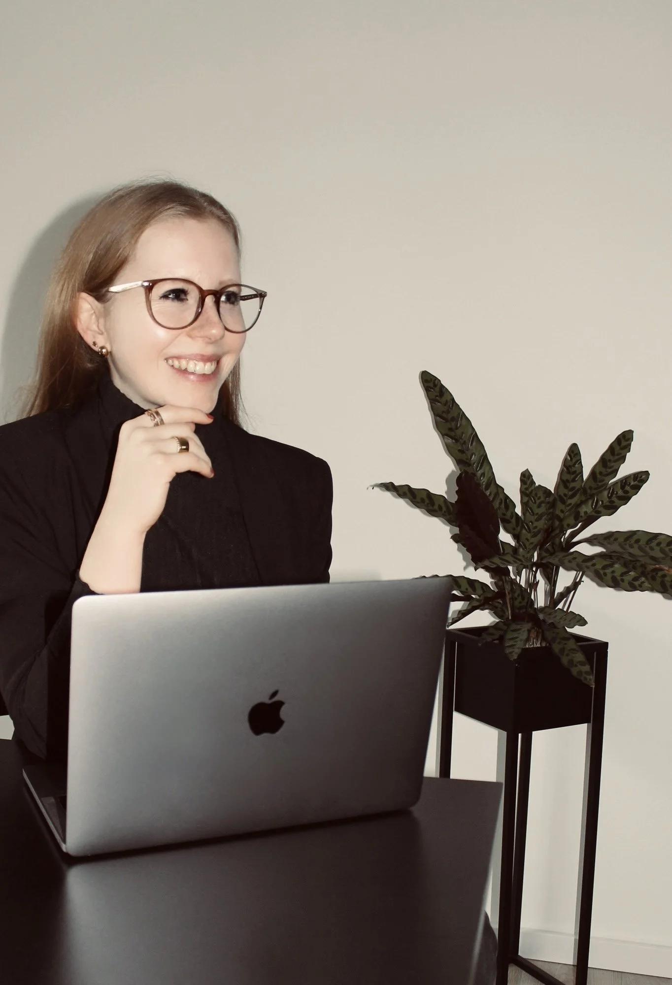 Eine lächelnde Frau mit Brille sitzt an einem schwarzen Tisch mit einem aufgeschlagenen Apple-Laptop. Im Hintergrund ist eine Zimmerpflanze in einem schwarzen Topf sichtbar.