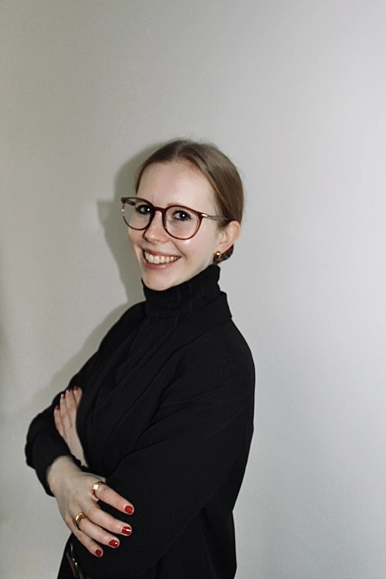 Junge Frau mit Brille, schwarzen Pullover und roten Nägeln steht vor weißer Wand, lächelt, hat kurz braune Haare und trägt goldene Ohrringe und Ringe.