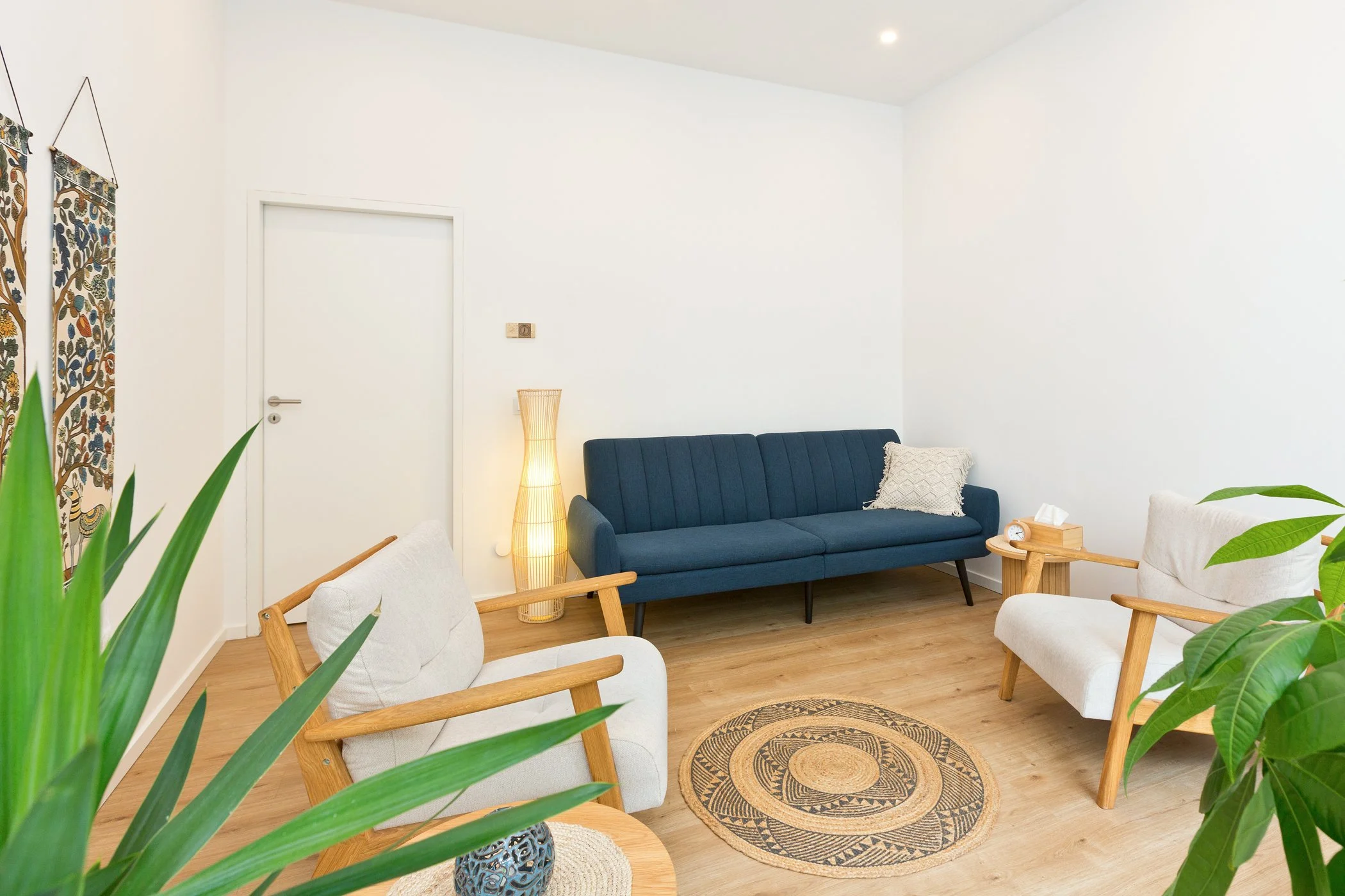 Heller Praxisraum mit blauer Couch, zwei weißen Sesseln, Juteteppich und Pflanzen