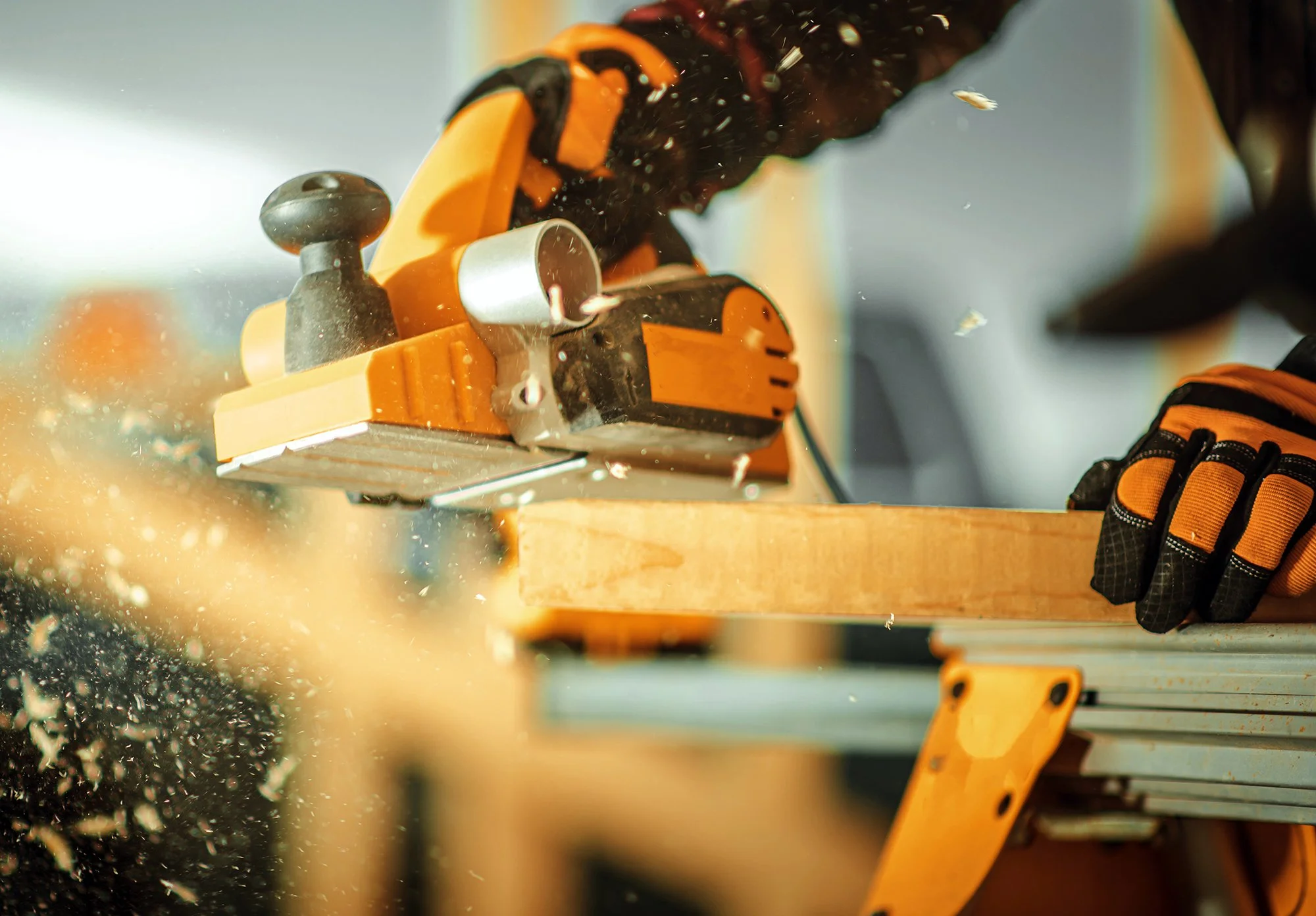 A person wearing gloves is using a yellow woodworking router to shape or carve wood on a workbench, with wood shavings flying.