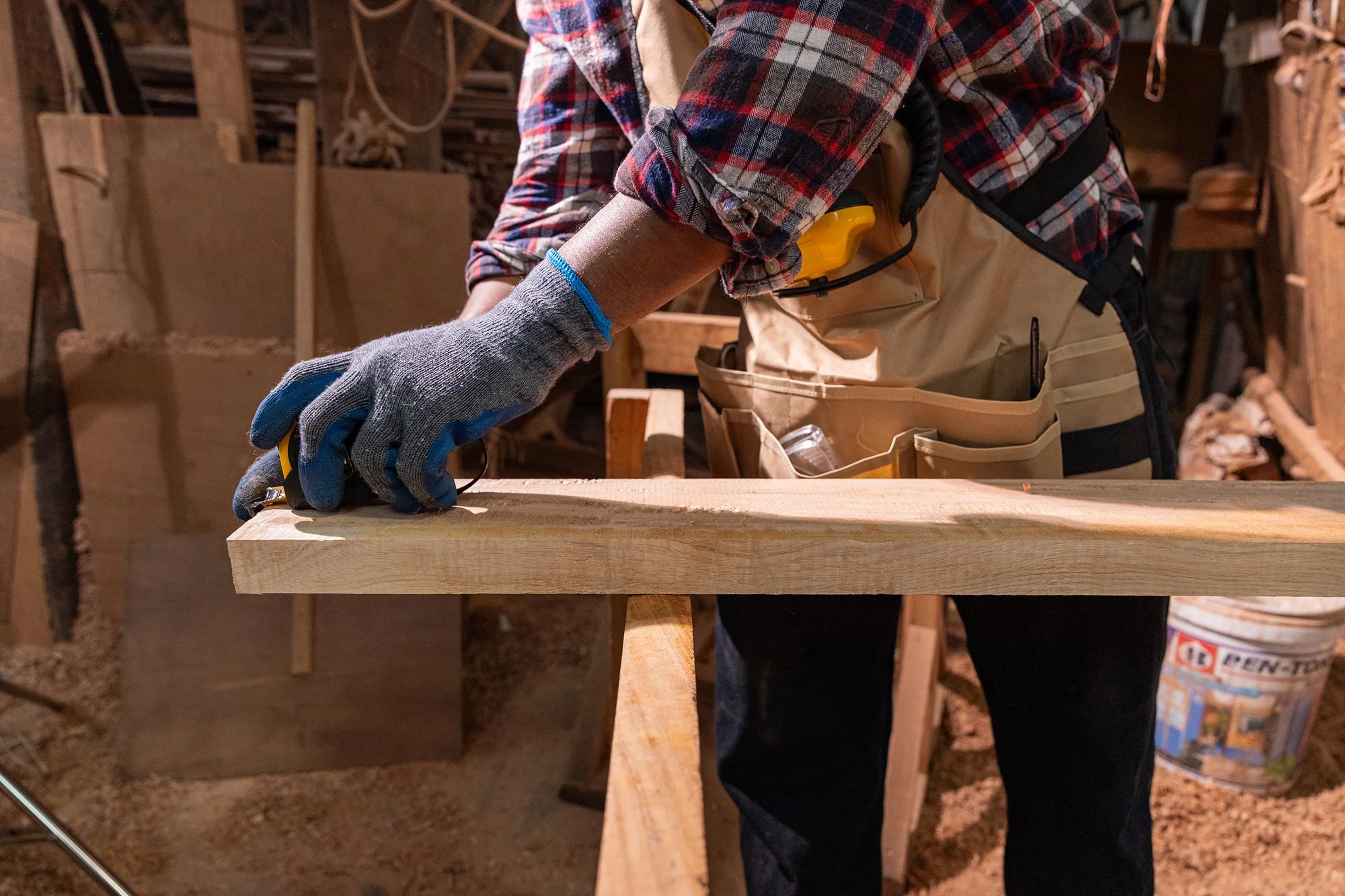 A person wearing a plaid shirt and tan apron working with a piece of wood in a woodworking shop. The person has a safety glove on one hand and is using a tape measure on the wood. The background shows woodworking tools and shelves.