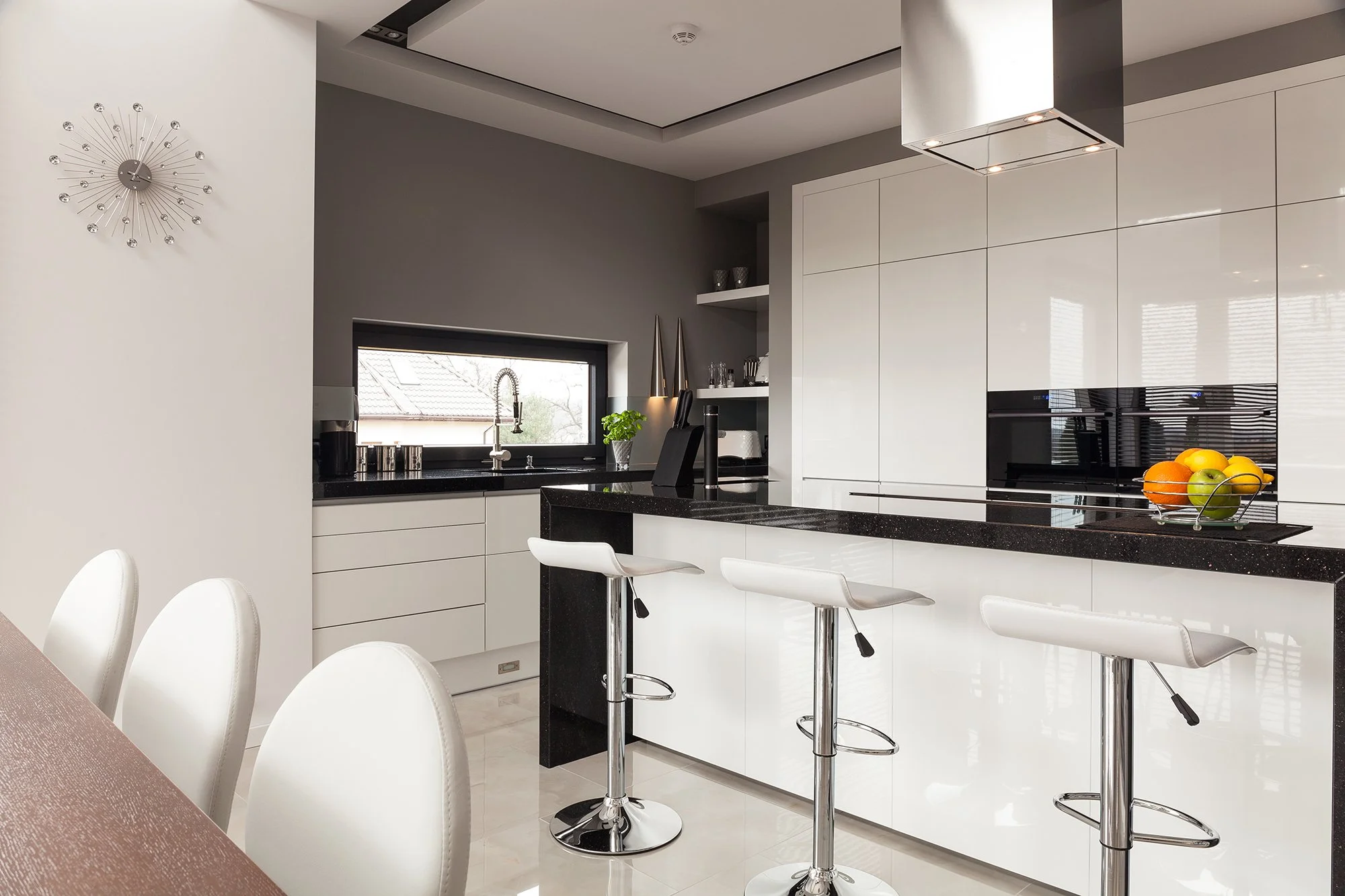 Modern kitchen with white cabinetry, black countertops, three white bar stools, a bowl of fruit, and a window with a view of neighboring rooftops.