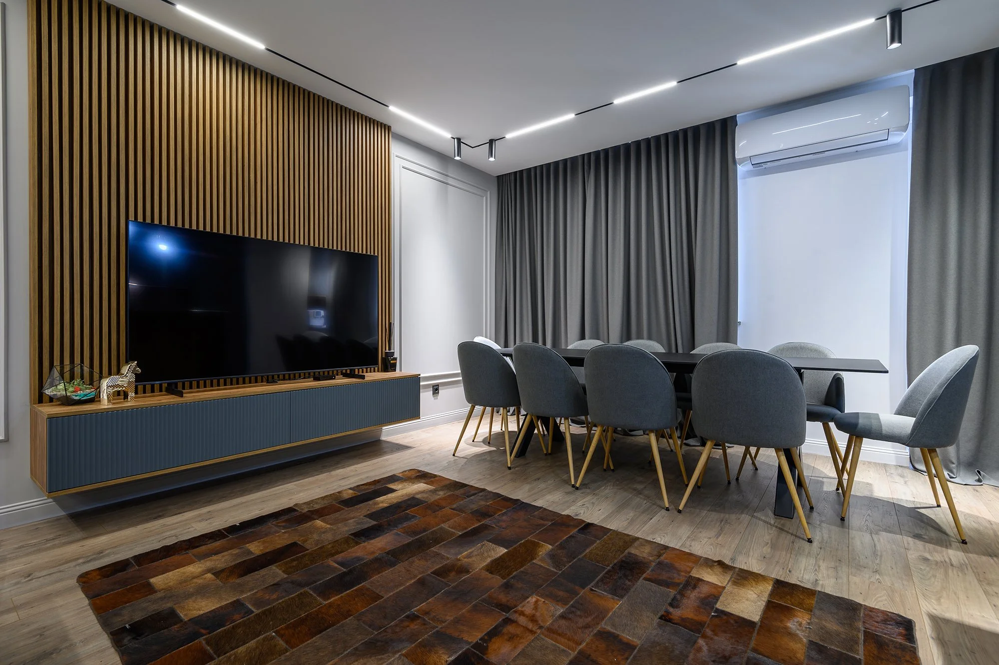 Modern dining room with a large rectangular table surrounded by ten grey chairs. A flat-screen TV is mounted on a wood-paneled accent wall. Grey curtains cover the window, with an air conditioner above. The floor is light wood, with a multicolored rug in the foreground.