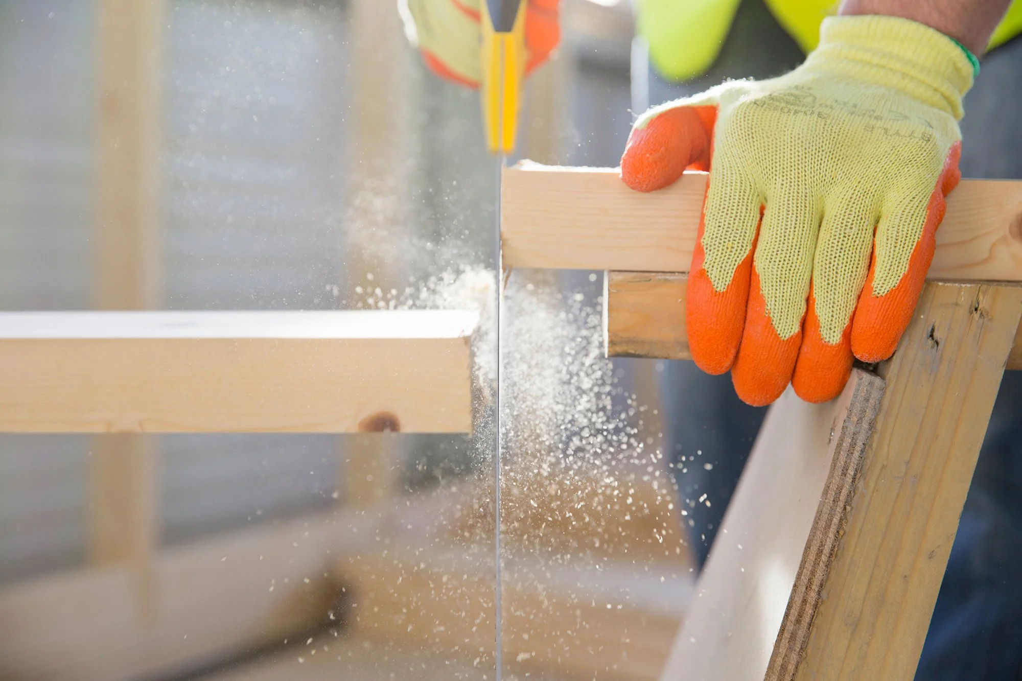 Close-up of a person's hand wearing yellow and orange work gloves, holding a wooden plank at an angle while cutting with a hand saw, with sawdust flying in the air.
