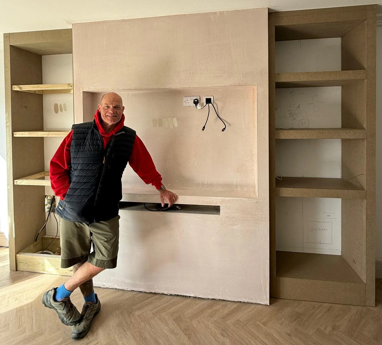 A man in outdoor clothing standing next to a partially constructed wooden built-in cabinet in a room under construction.