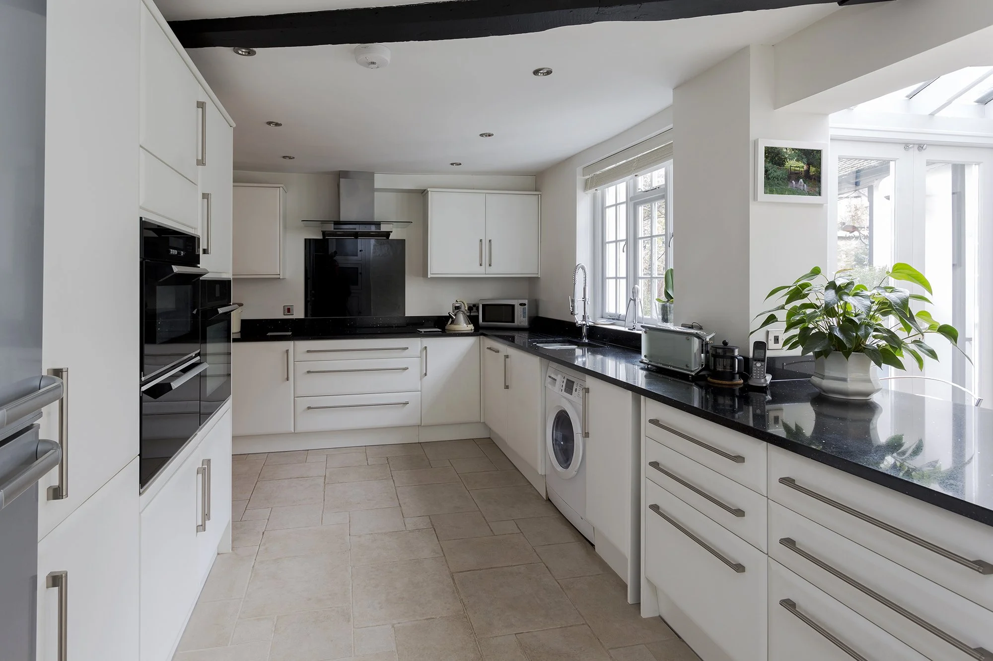 Modern white kitchen with black countertops, appliances including oven, microwave, washing machine, and small plants near windows.