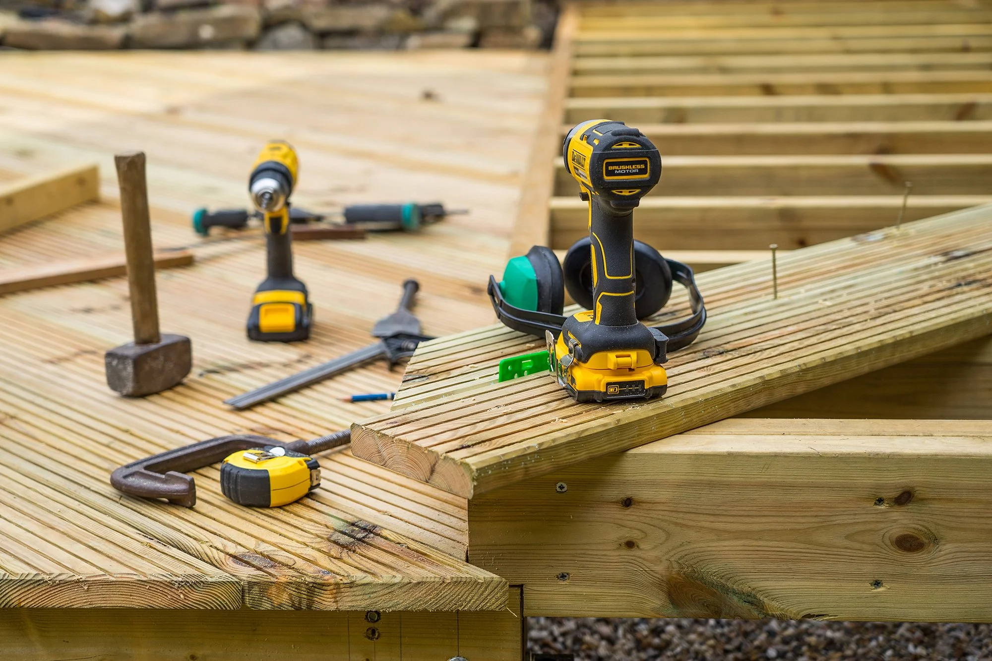 Construction tools including a cordless drill with a yellow and black handle, a measuring tape, a C-clamp, a square ruler, and a hammer are arranged on wooden planks at a construction site.