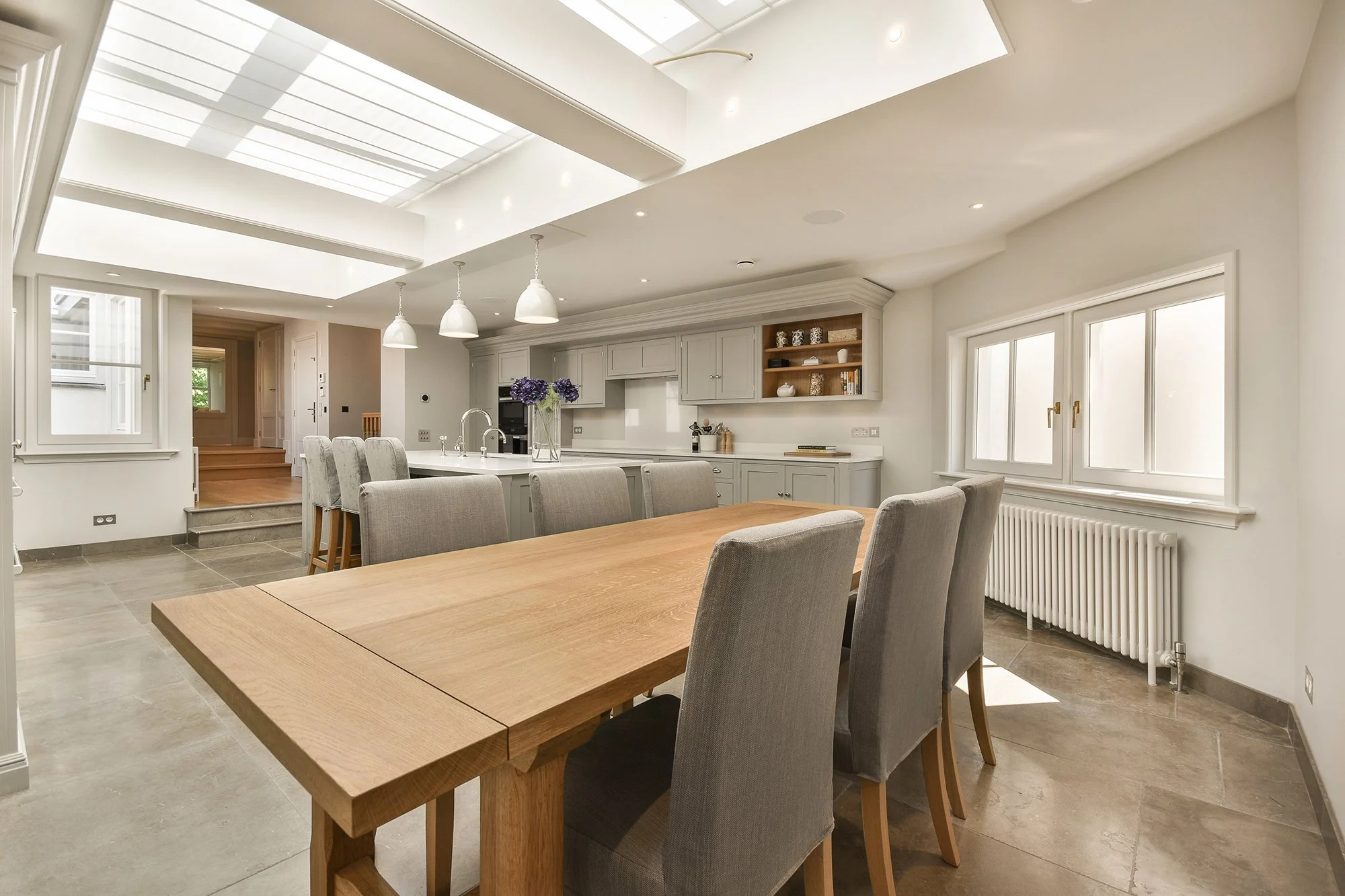 Bright, spacious kitchen and dining area with skylight, white cabinetry, wooden dining table, and gray upholstered chairs.