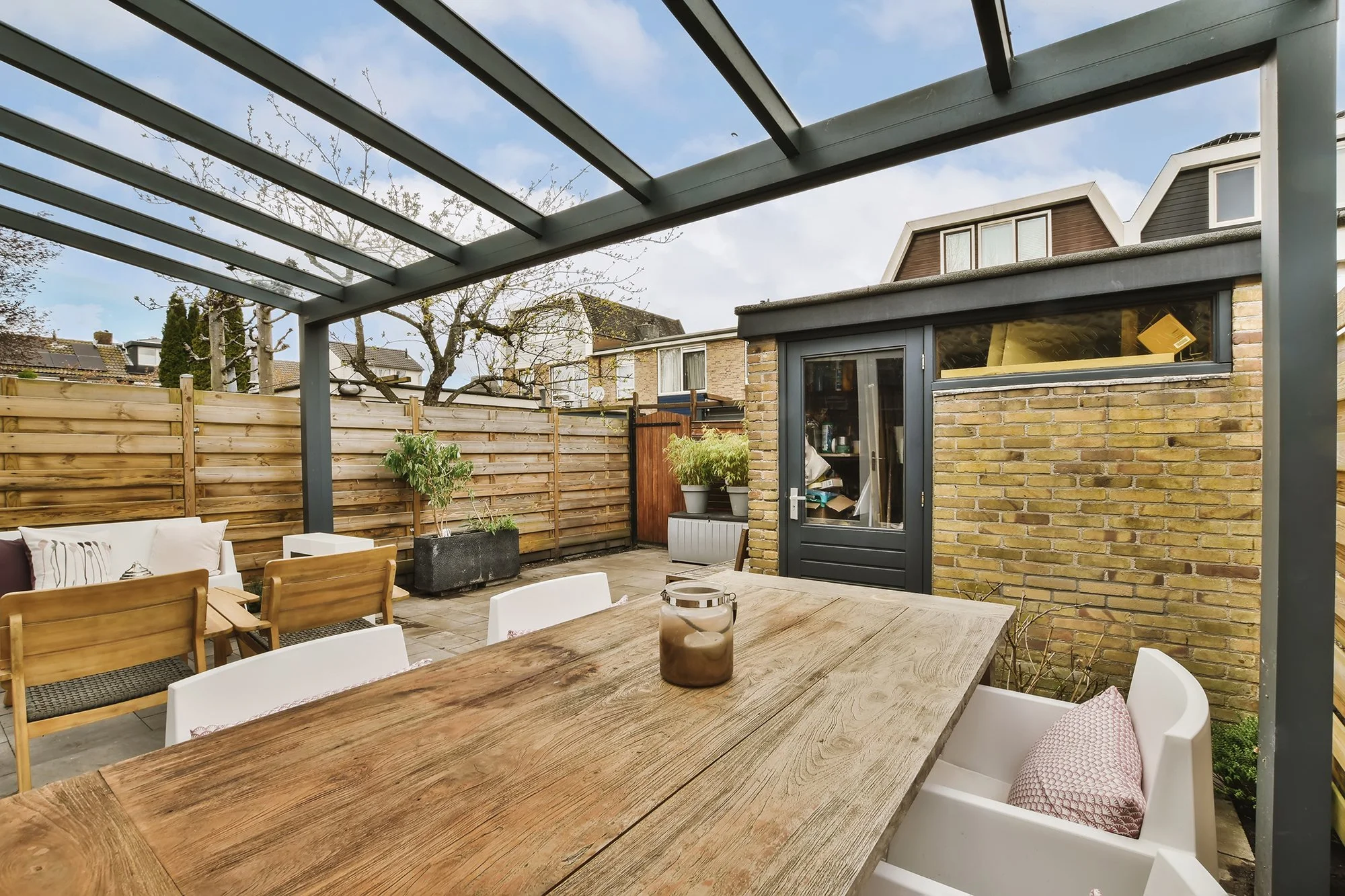 Outdoor patio with a wooden dining table, chairs, potted plants, and a brick shed, enclosed by a wooden fence and a glass canopy overhead.