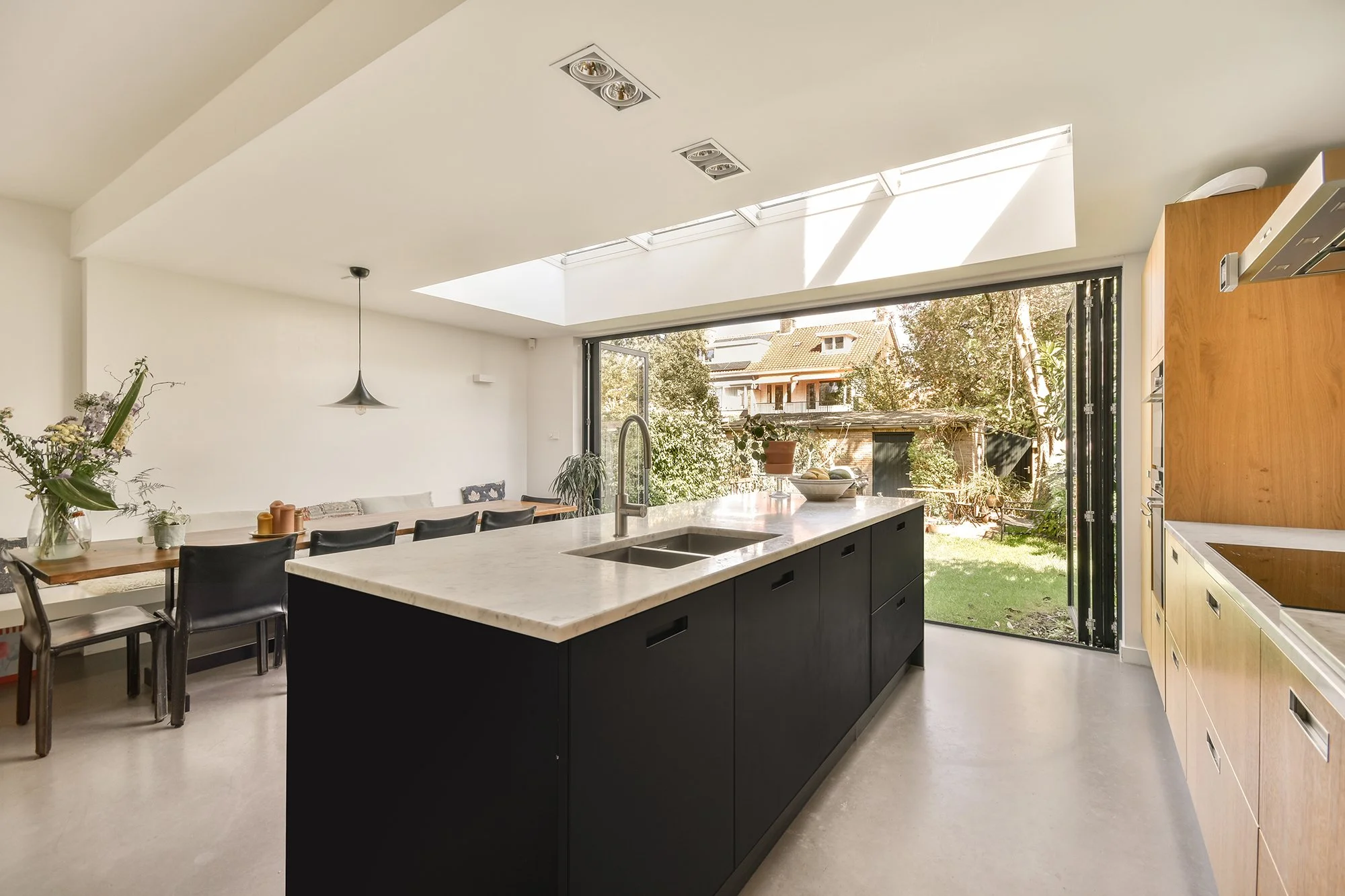Modern kitchen with black island, wooden cabinets, open view to garden, skylight, and sliding glass door.