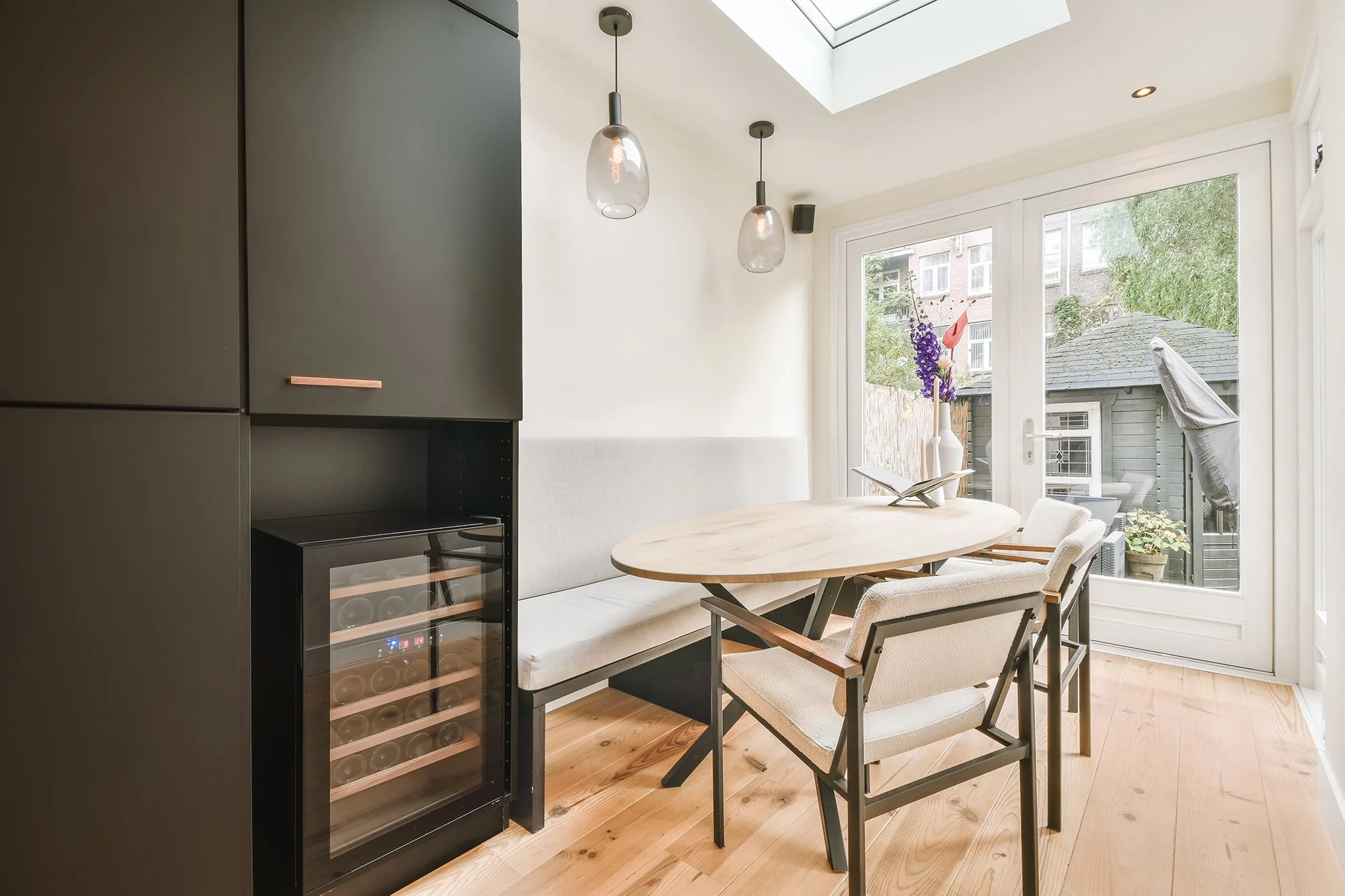 Dining area with a wooden table, an upholstered bench and chairs, a black wine cooler, and large glass doors leading outside, with ceiling pendant lights and a skylight.