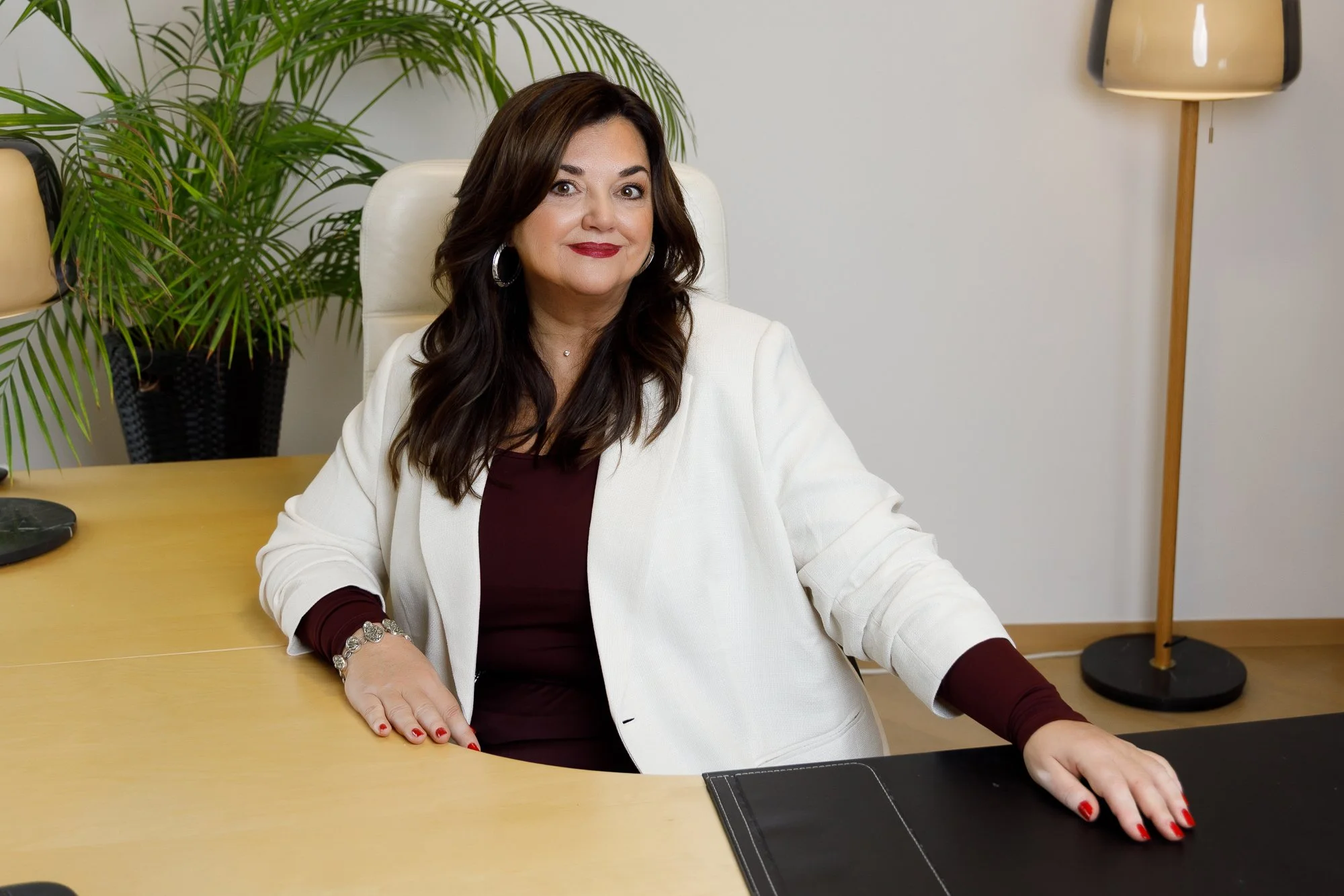 Frau sitzt an einem Büro-Schreibtisch mit einem weißen Mantel, roten Kleid, dunklen Haaren, rote Nägel, Schuhe. Im Hintergrund eine Zimmerpflanze und eine Stehlampe.