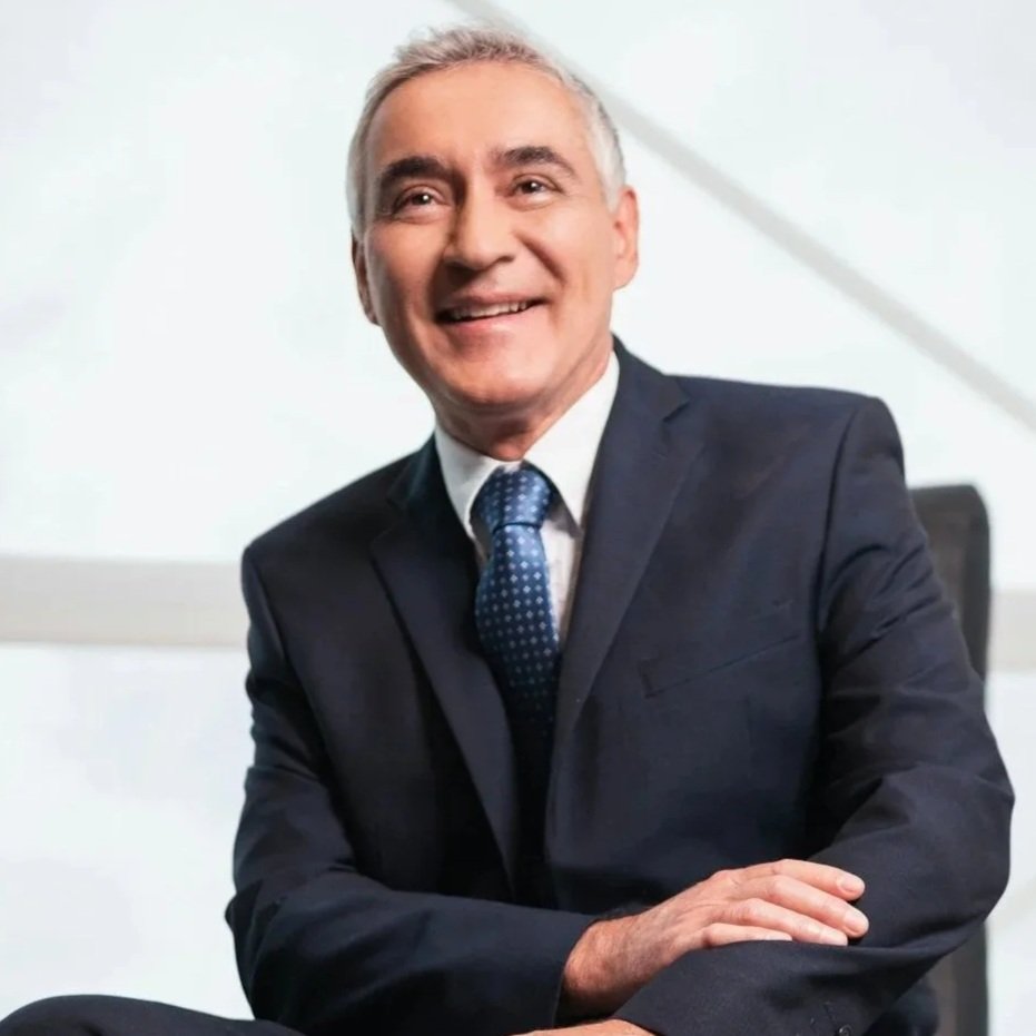 Ein Mann in einem dunklen Anzug und einer blauen Krawatte, der lächelnd vor weißem Hintergrund sitzt.