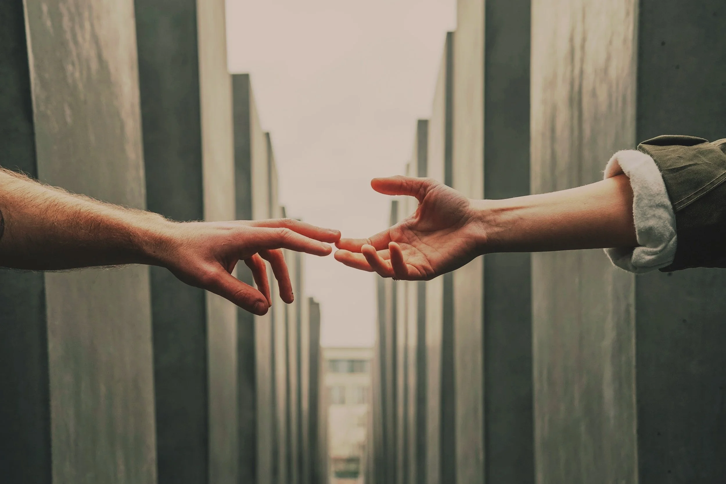 Two hands reaching towards each other with index fingers almost touching, in an outdoor setting between tall structures.