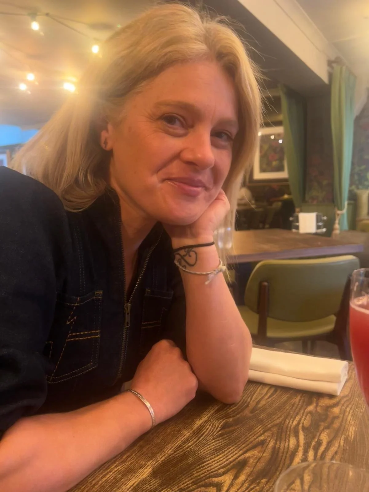 A woman with blonde hair smiling, resting her chin on her hand, sitting at a wooden table in a restaurant or cafe with warm lighting and green curtains.
